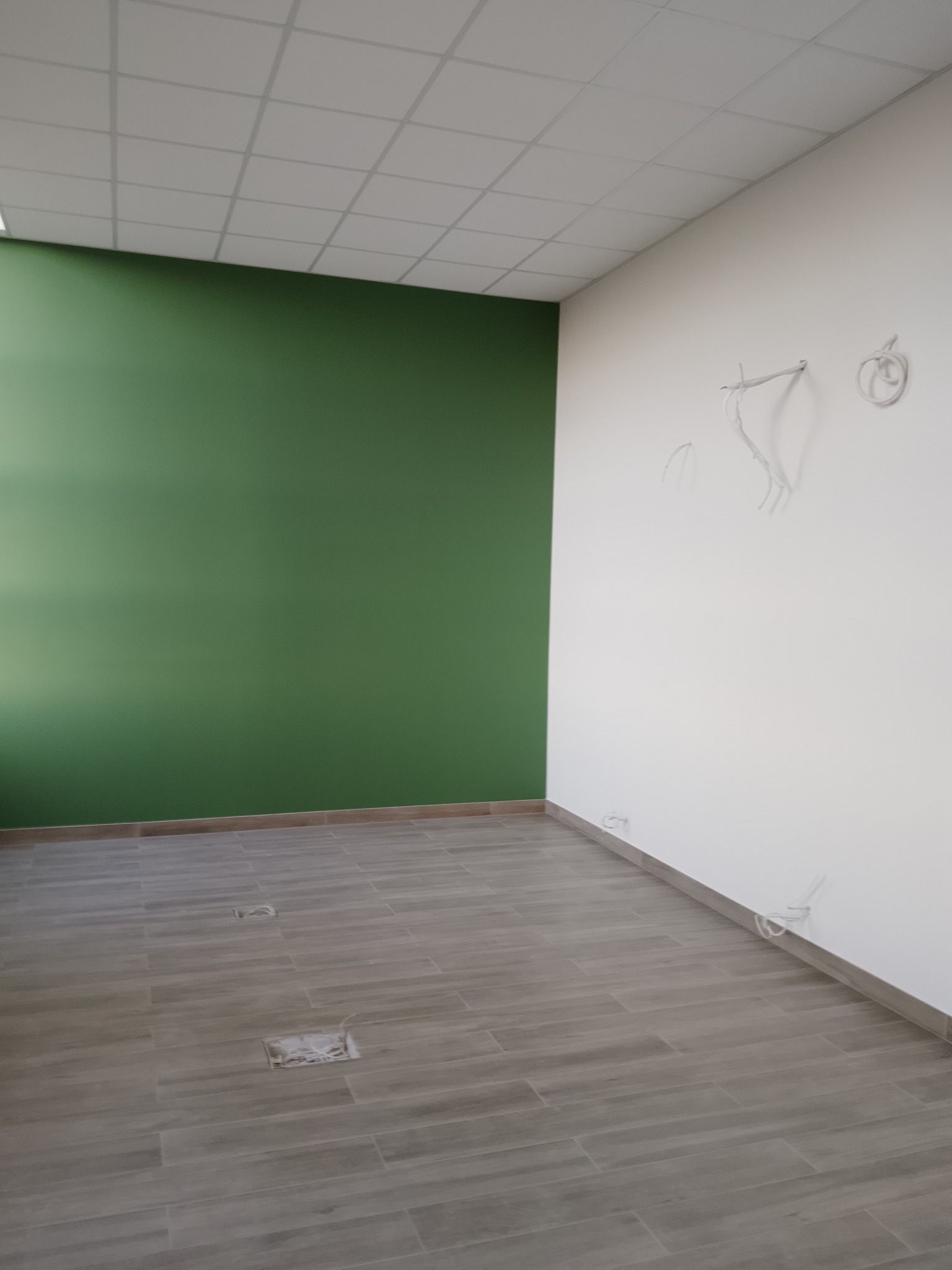 Wnętrze biura po remoncie: zielona ściana, szare płytki podłogowe, biała ściana z wystającymi przewodami. Widoczne otwory w podłodze. Sufit kasetonowy.