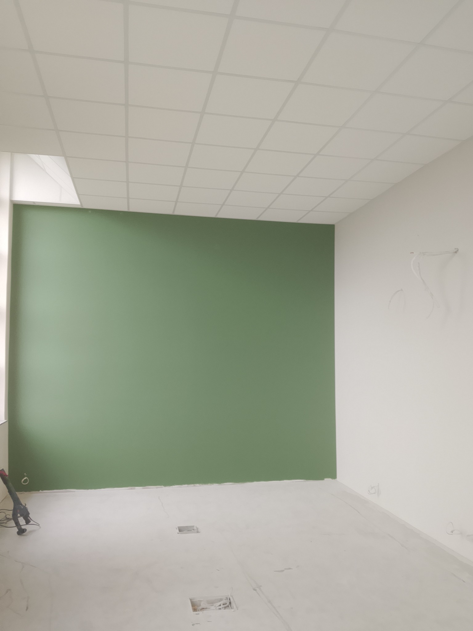Wnętrze w trakcie prac: zielona ściana, sufit kasetonowy, szpachlowana podłoga z widocznymi otworami. W rogu narzędzie do szlifowania. Widoczne przewody elektryczne.
