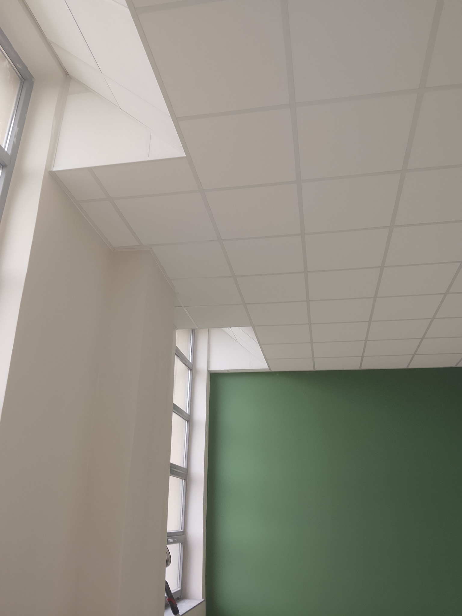 Kwadratowa, biała podwieszana sufitowa konstrukcja z widoczną zieloną ścianą i oknem. Ujęcie z dołu, ukazujące detale łączeń i fakturę sufitu.