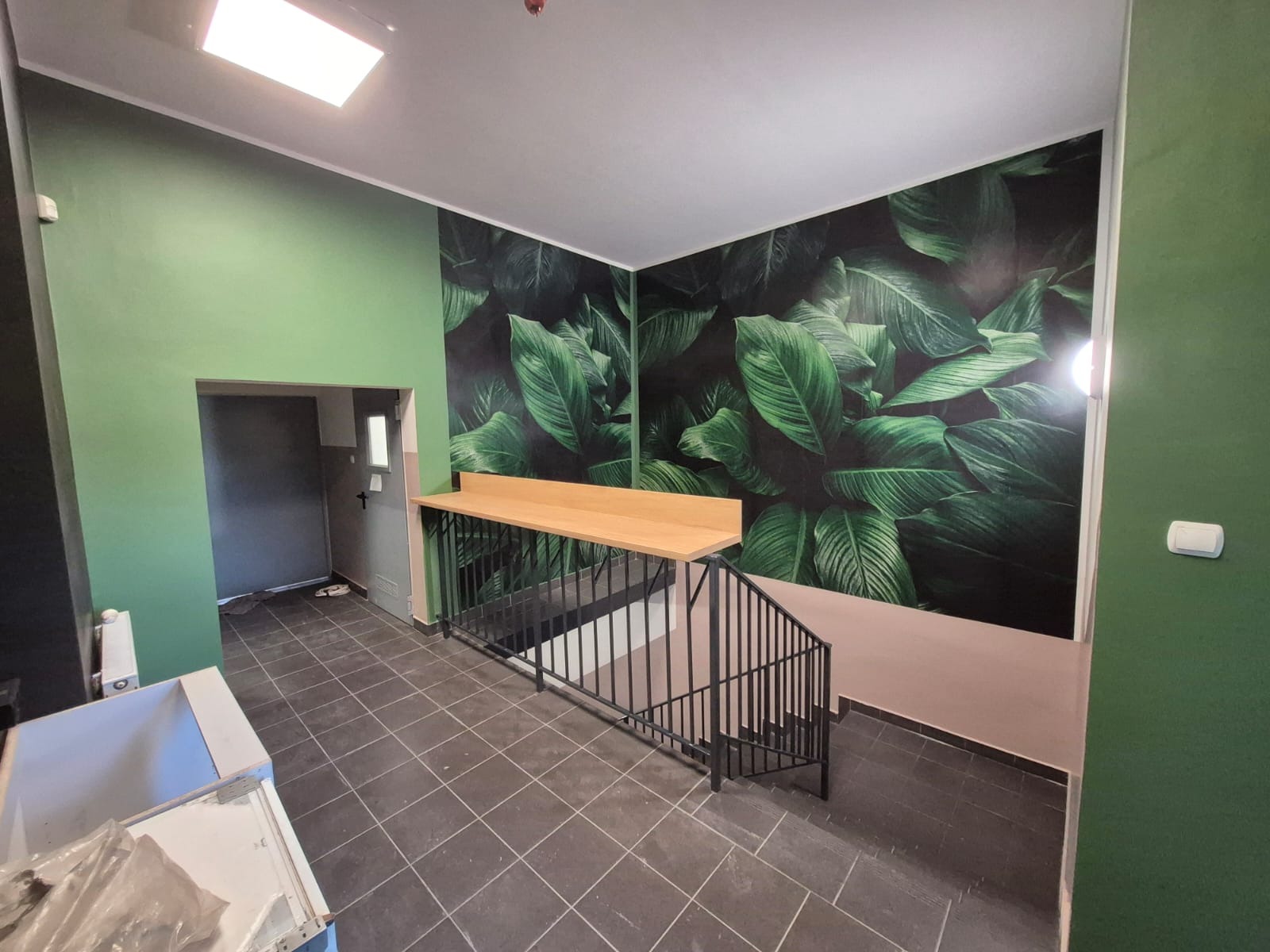 Wnętrze po odświeżeniu: zielone ściany, fototapeta z motywem liści, drewniany blat nad schodami z czarną balustradą i szara podłoga z płytek. Widoczny fragment sprzętu.