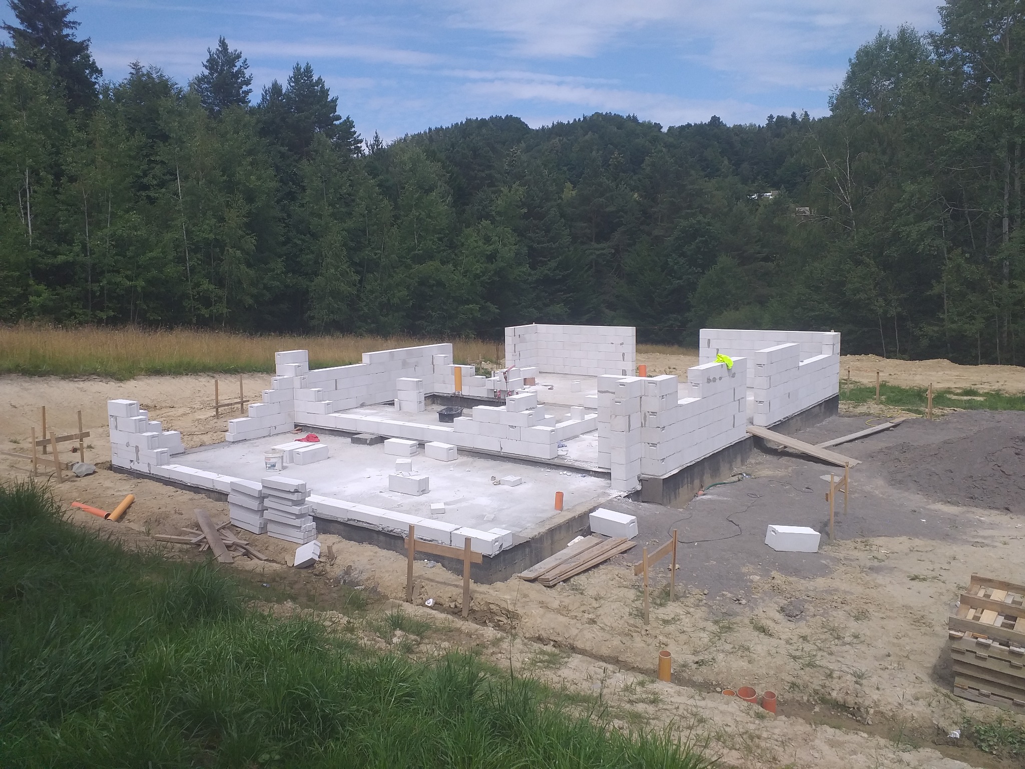 Wznoszenie fundamentów z bloczków betonu komórkowego na betonowej płycie. Widoczne zbrojenia i elementy instalacji. W tle las i błękitne niebo.