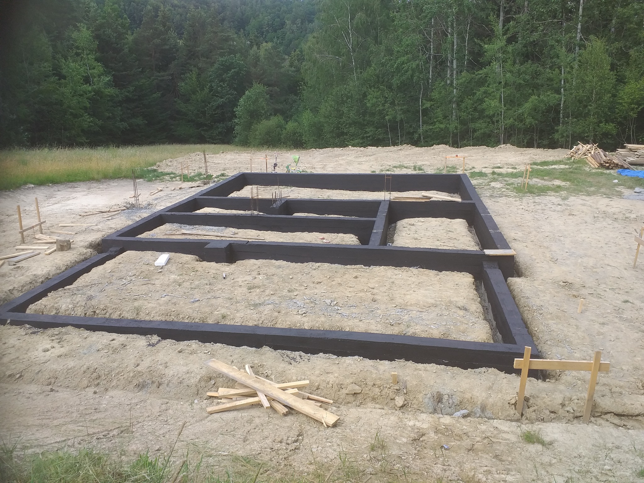 Zabezpieczone, pomalowane fundamenty domu jednorodzinnego na działce z widokiem na las. Widoczne szalunki i wystające pręty zbrojeniowe.