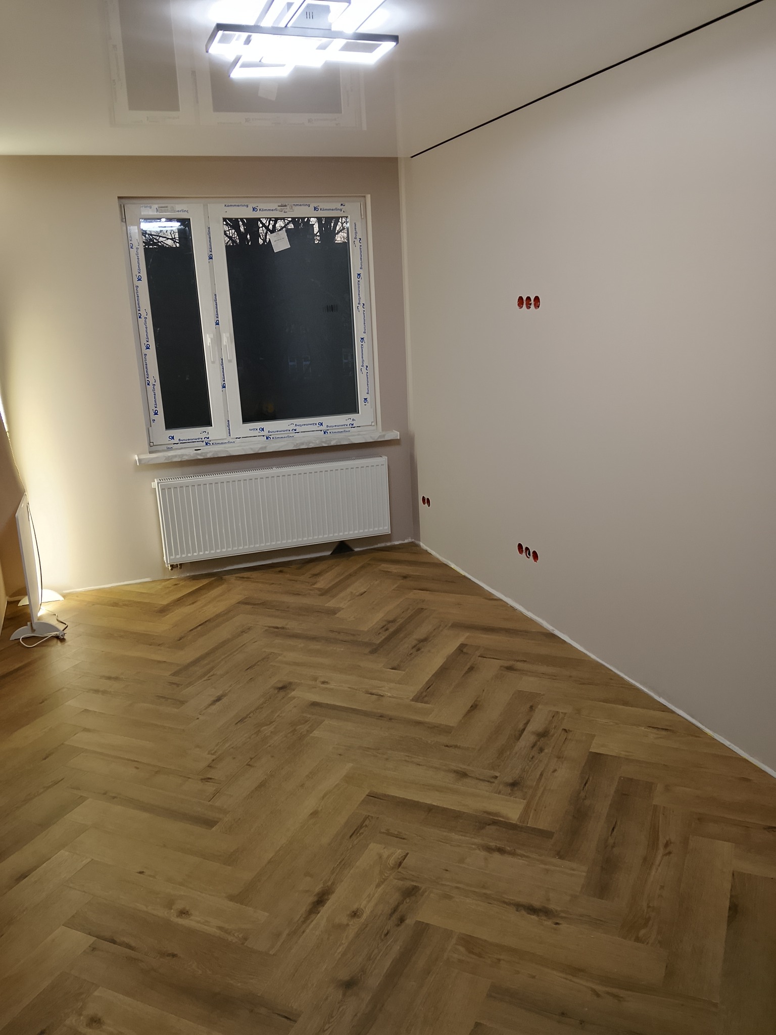 Jasne panele ułożone w jodełkę w pokoju z oknem, grzejnikiem i gładkimi ścianami z widocznymi puszkami elektrycznymi. Nowoczesne oświetlenie sufitowe.