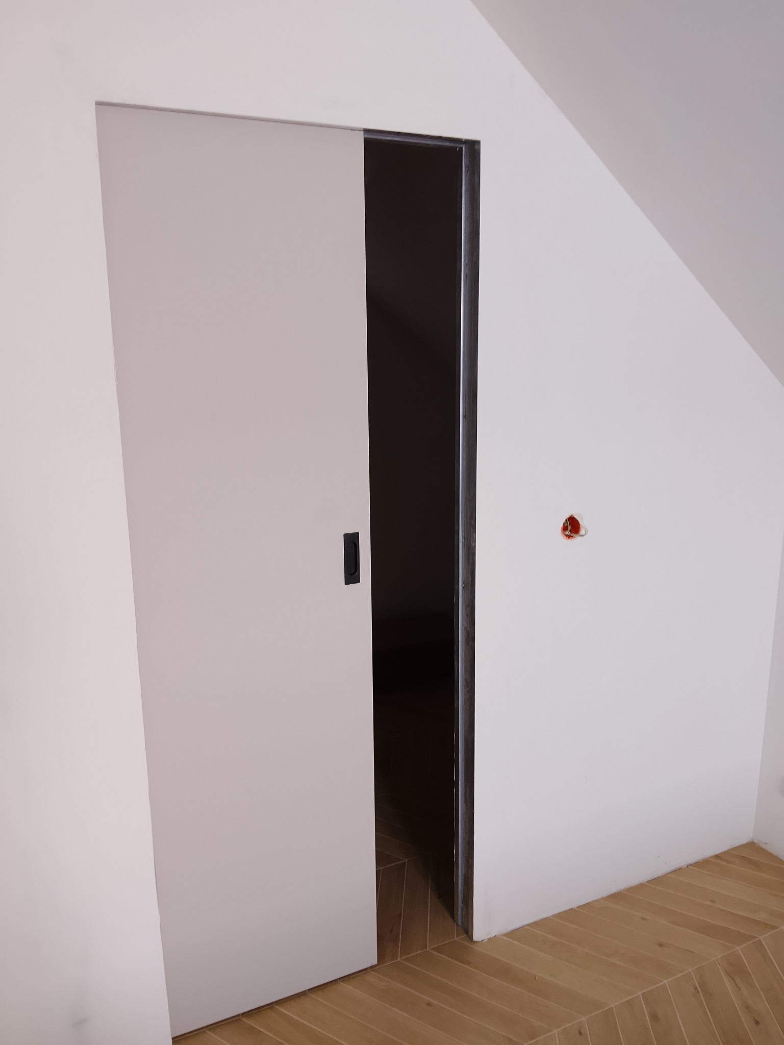 Białe, przesuwne drzwi w skosie poddasza, z widoczną czarną klamką i fragmentem drewnianej podłogi ułożonej w jodełkę. Na ścianie widoczne gniazdko elektryczne.