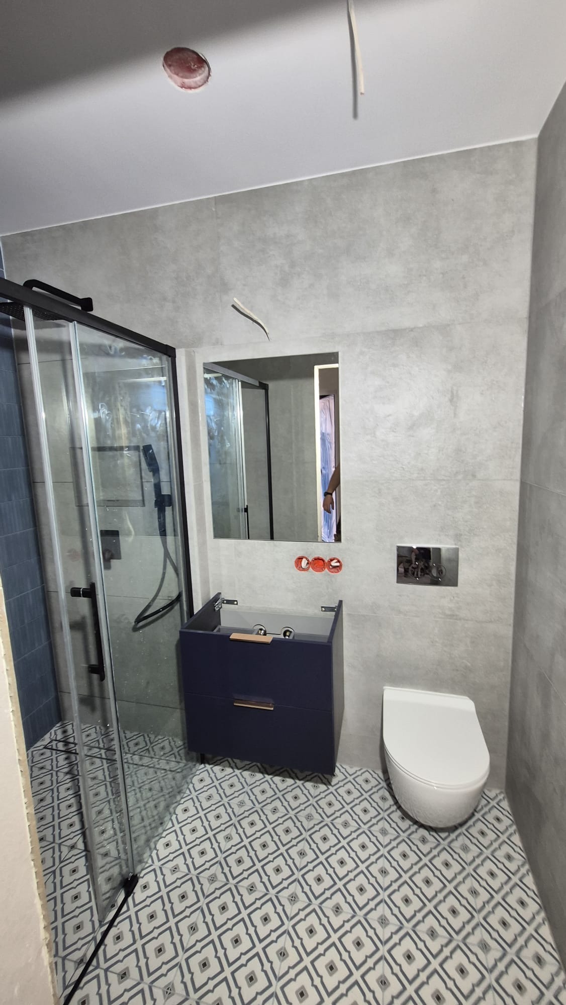 Nowoczesna łazienka w szarościach z kabiną prysznicową, umywalką w szafce i toaletą. Wzór na podłodze dodaje charakteru. Widoczne elementy instalacji.