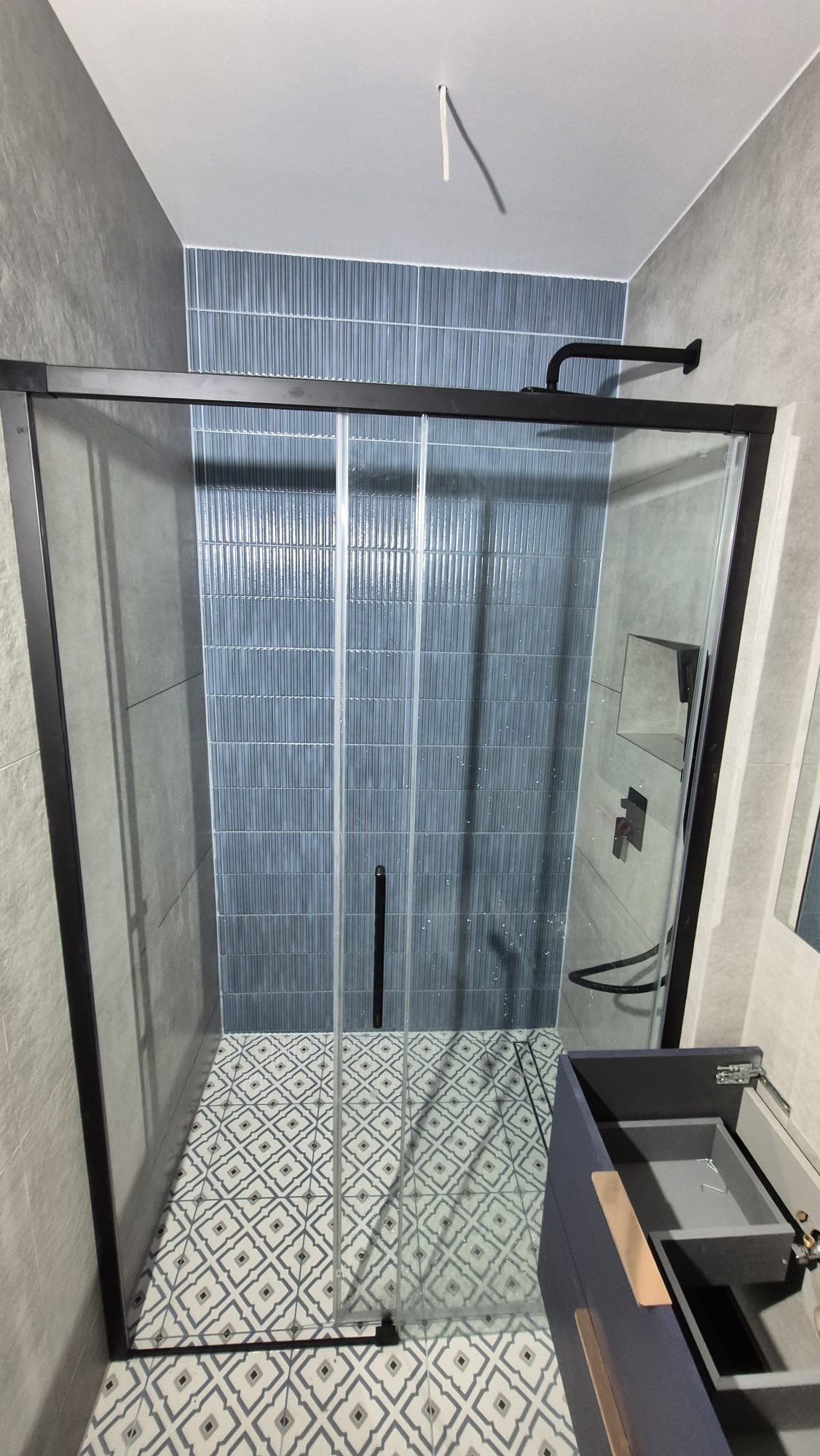 Nowoczesna kabina prysznicowa z czarną ramą, błękitne płytki na ścianie, wzorzysta podłoga. Widoczna umywalka z szafką w odcieniach szarości. Minimalistyczny design.