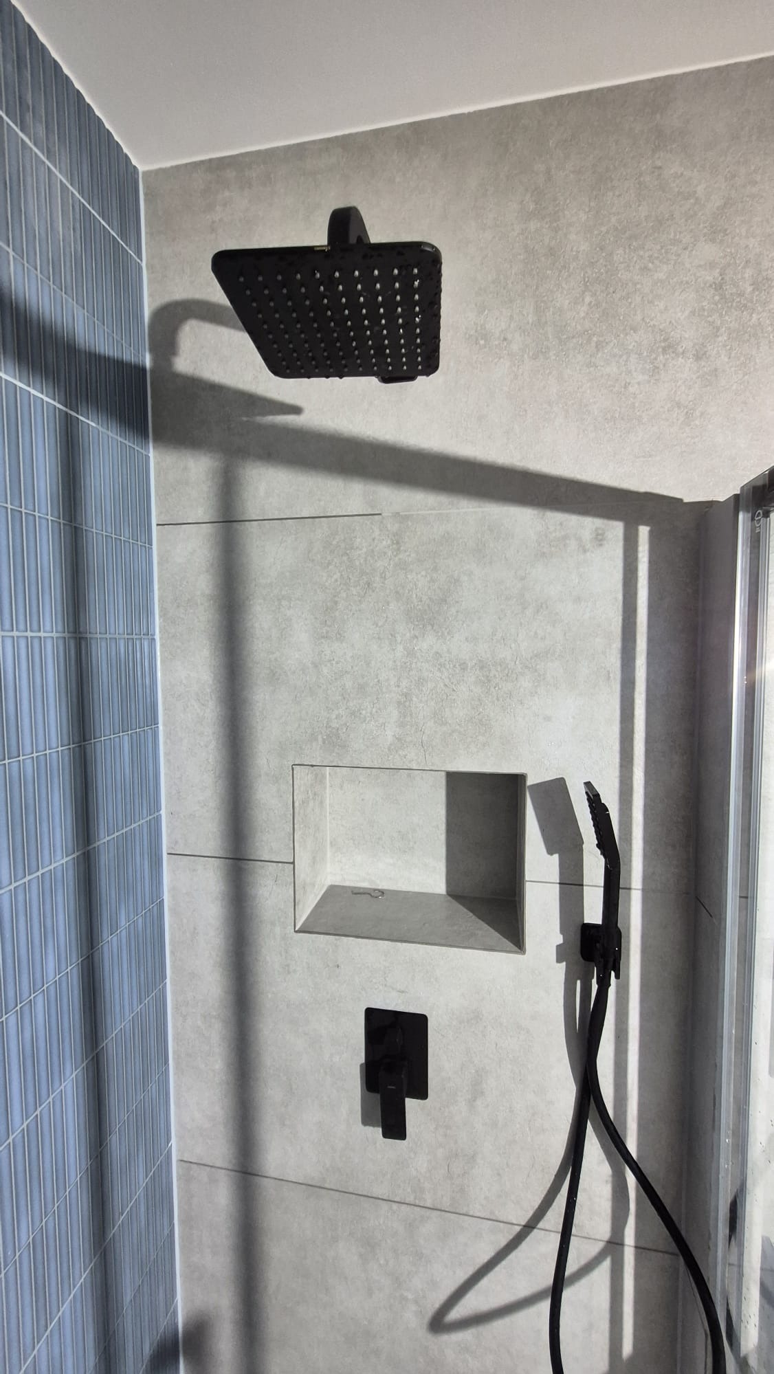 Nowoczesna kabina prysznicowa z czarną armaturą: deszczownica, bateria podtynkowa i wnęka w szarej płytce. Akcent stanowi ściana z niebieskiej, wąskiej glazury.