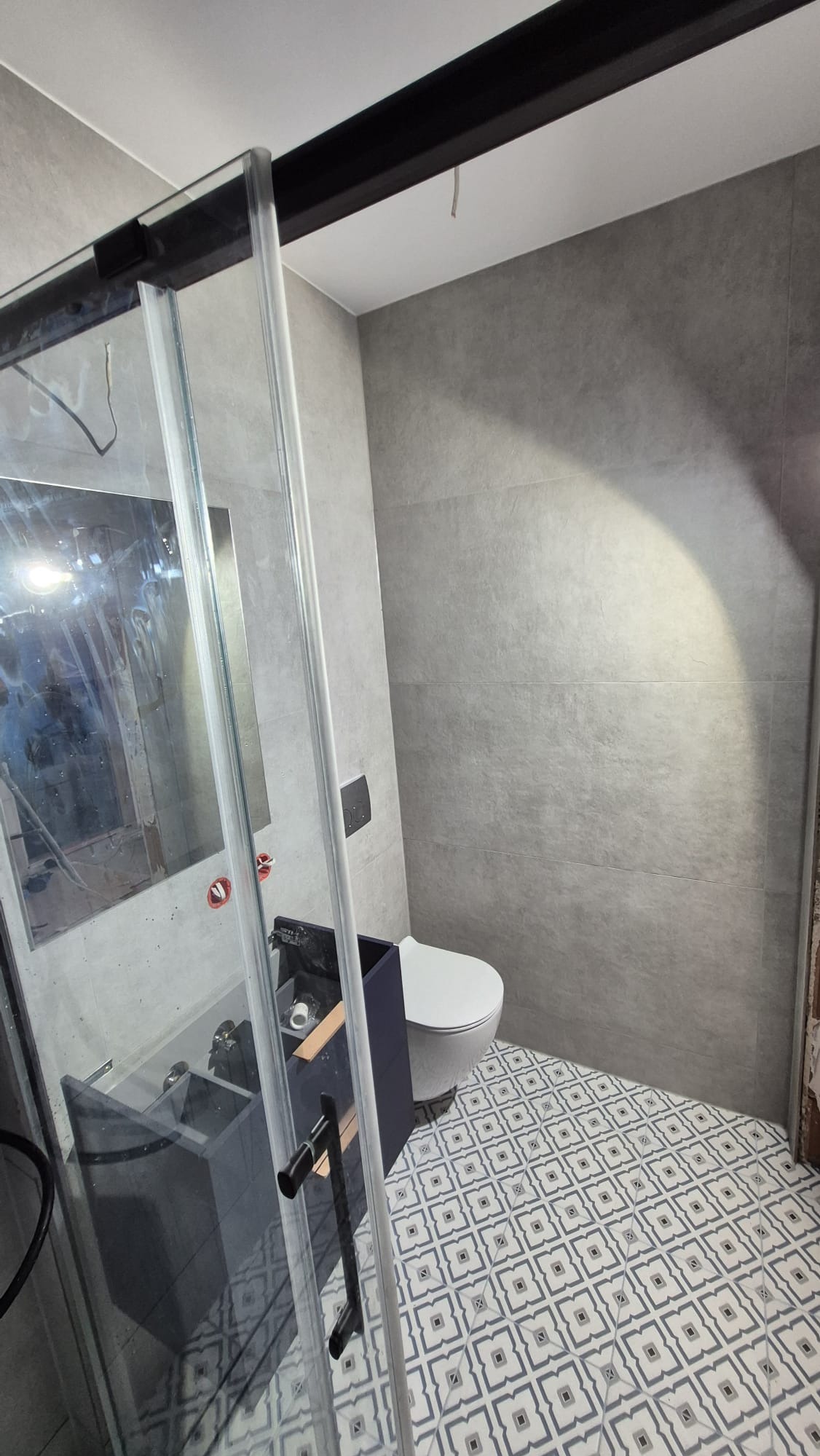 Wykończona łazienka z szarą glazurą, wzorzystą podłogą i kabiną prysznicową z czarnymi profilami. Widoczna toaleta i szafka pod umywalkę w kolorze granatowym.