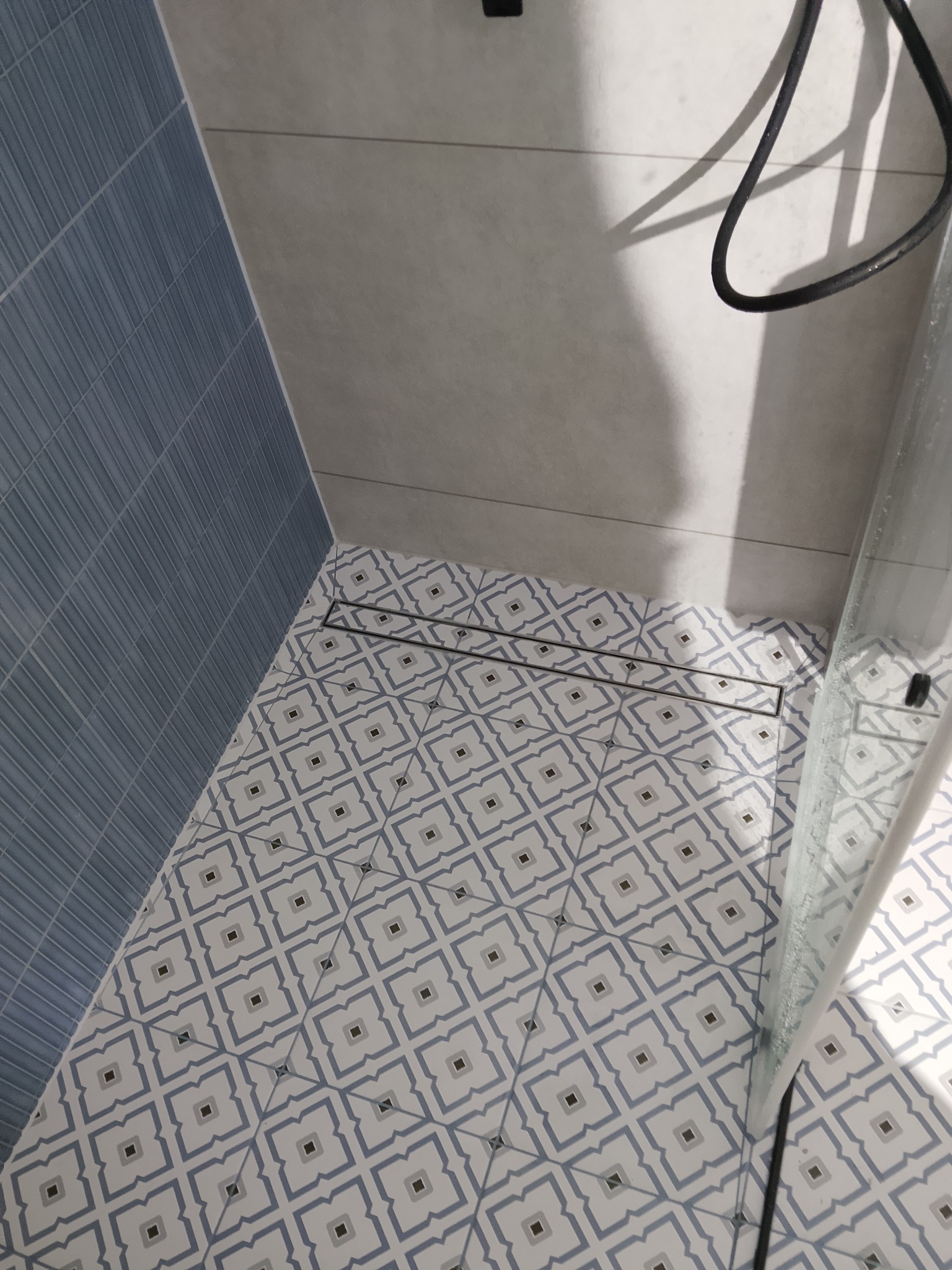 Wykończona łazienka z niebieskimi płytkami na ścianie i wzorzystą podłogą. Widoczny odpływ liniowy i fragment kabiny prysznicowej. Ujęcie z góry.