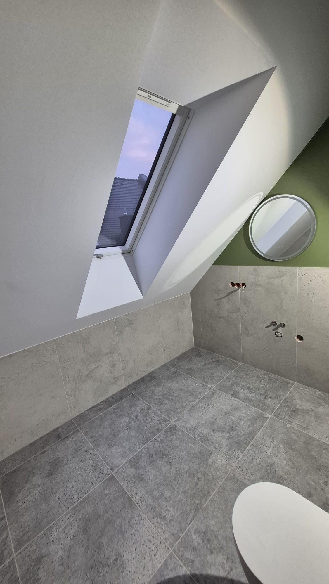 Szara łazienka na poddaszu z oknem dachowym, płytkami imitującymi kamień i okrągłym lustrem. Widoczne wyprowadzenia instalacji. Ściana akcentowa w kolorze zielonym.