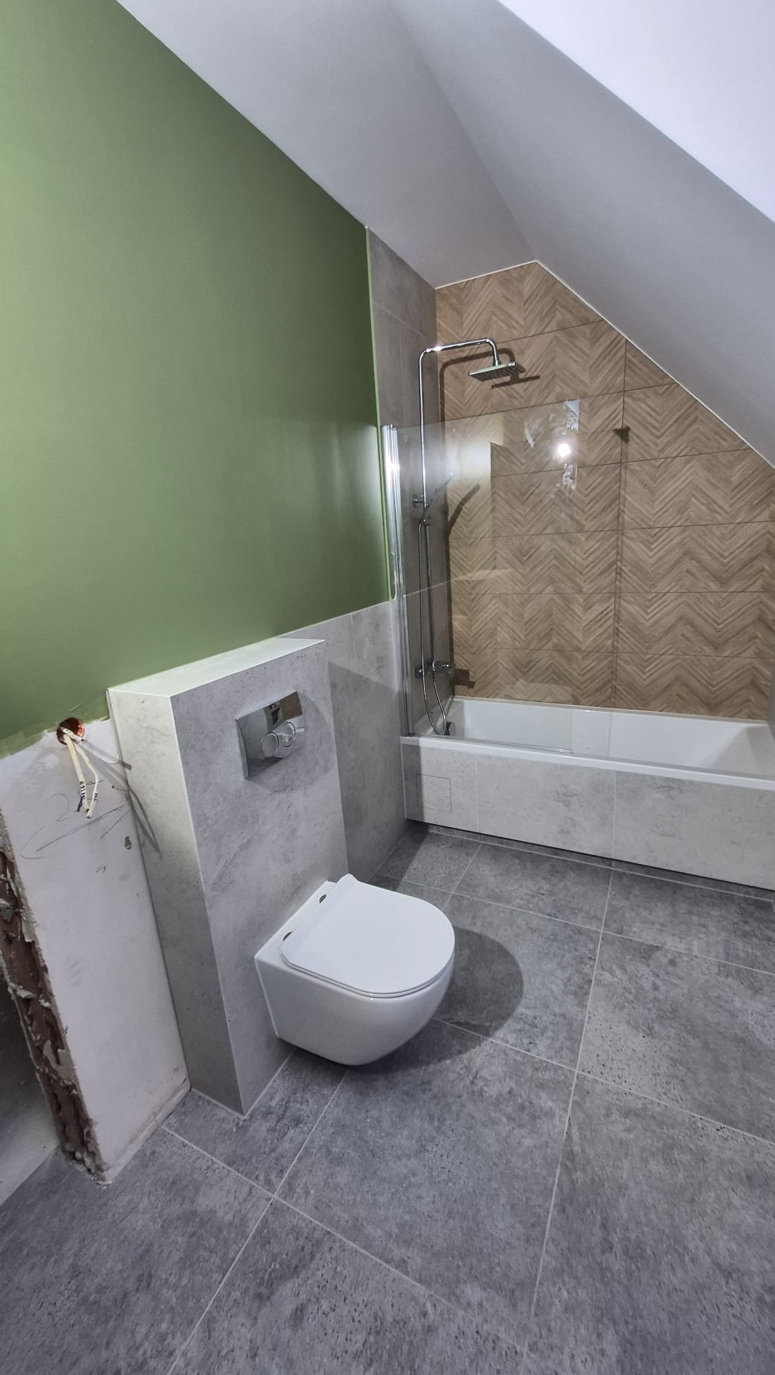 Wykończona łazienka na poddaszu z szarą podłogą i częściowo zieloną ścianą. Widoczna toaleta podwieszana, wanna z parawanem i płytki imitujące drewno na ścianie.