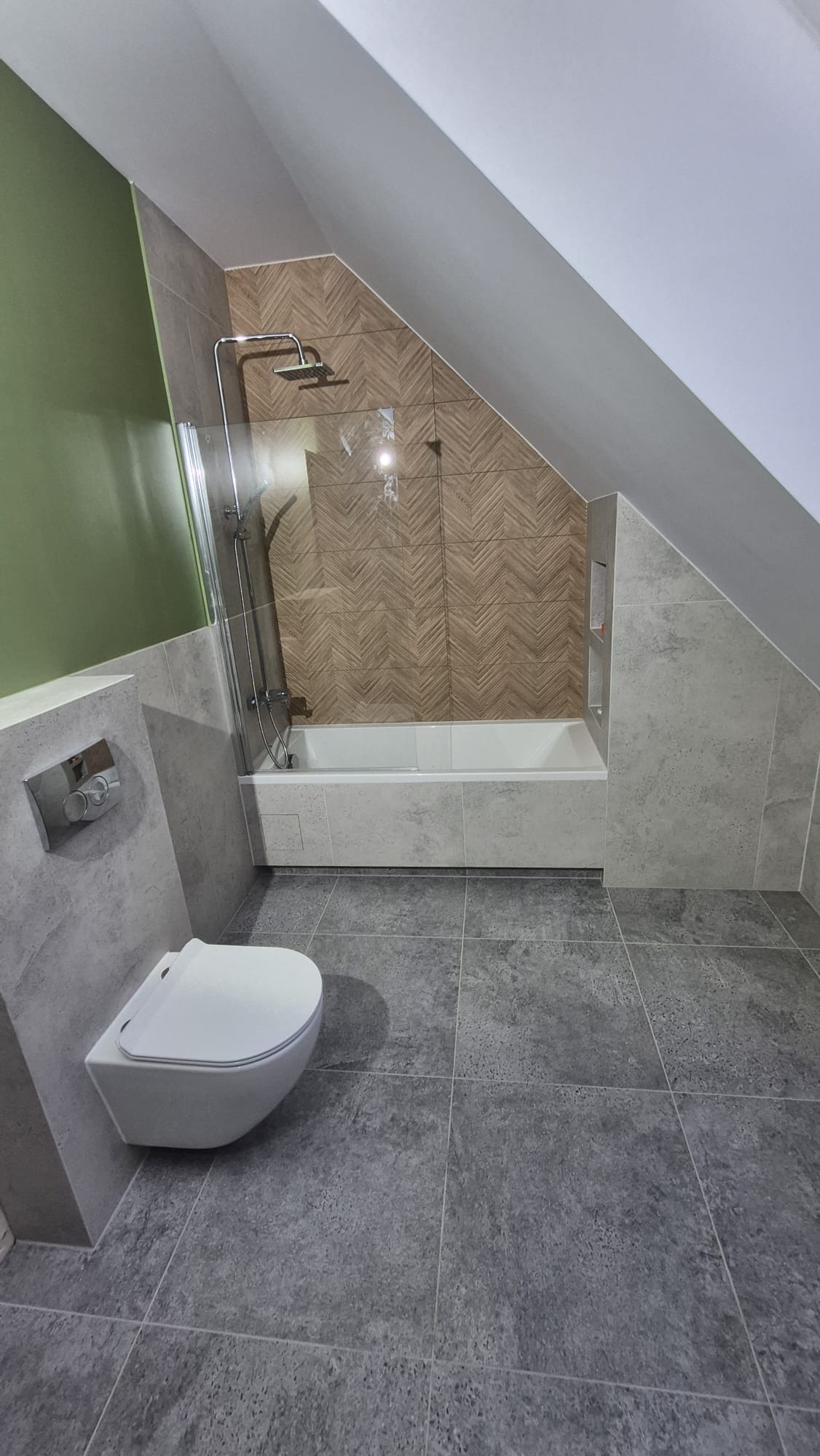 Nowoczesna łazienka na poddaszu z szarą glazurą, białą wanną i podwieszaną toaletą. Uwagę przykuwa ściana nad wanną wykończona płytkami w jodełkę.