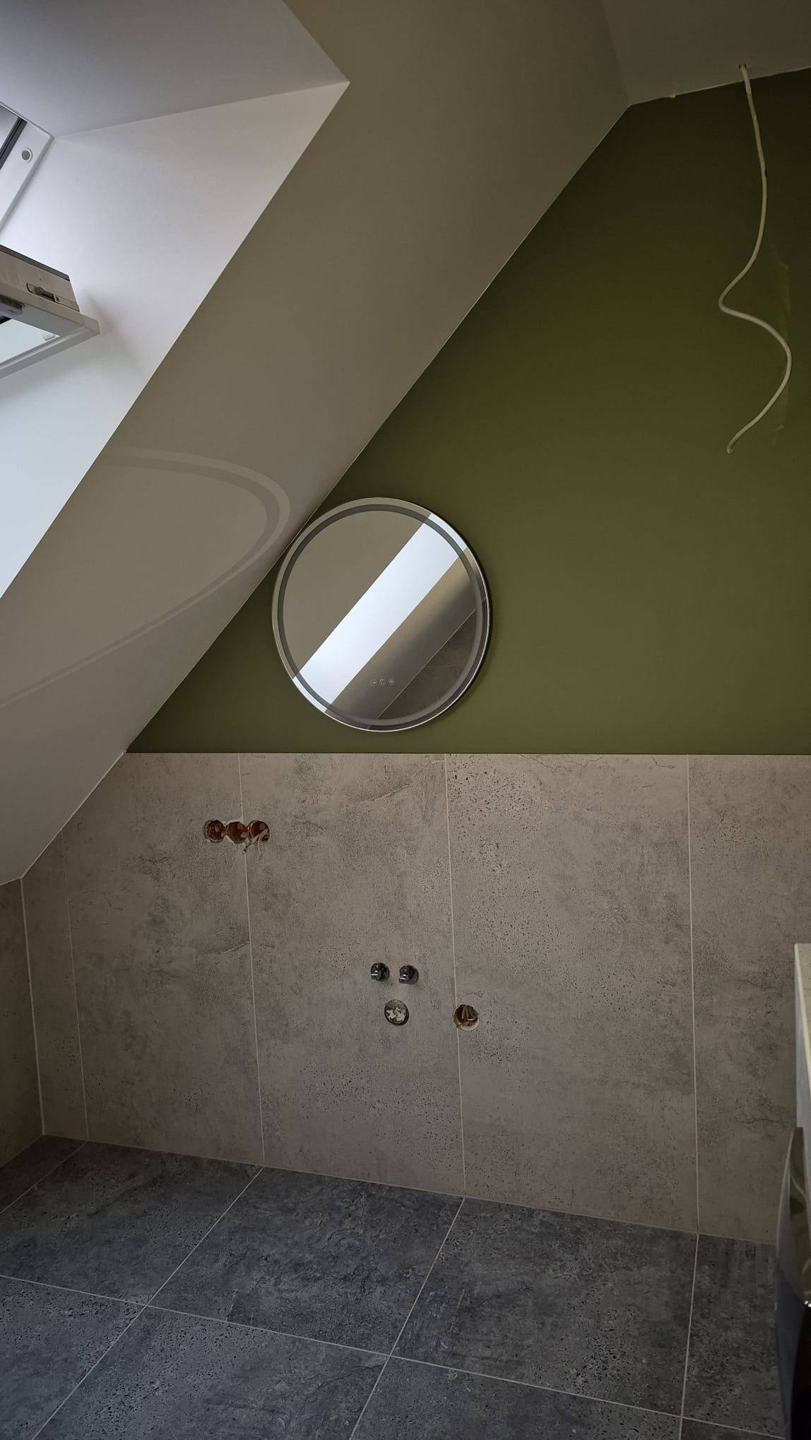 Wykończona łazienka na poddaszu: szare płytki na ścianach i podłodze, okrągłe lustro z podświetleniem na oliwkowej ścianie. Widoczne wyprowadzenia instalacji.