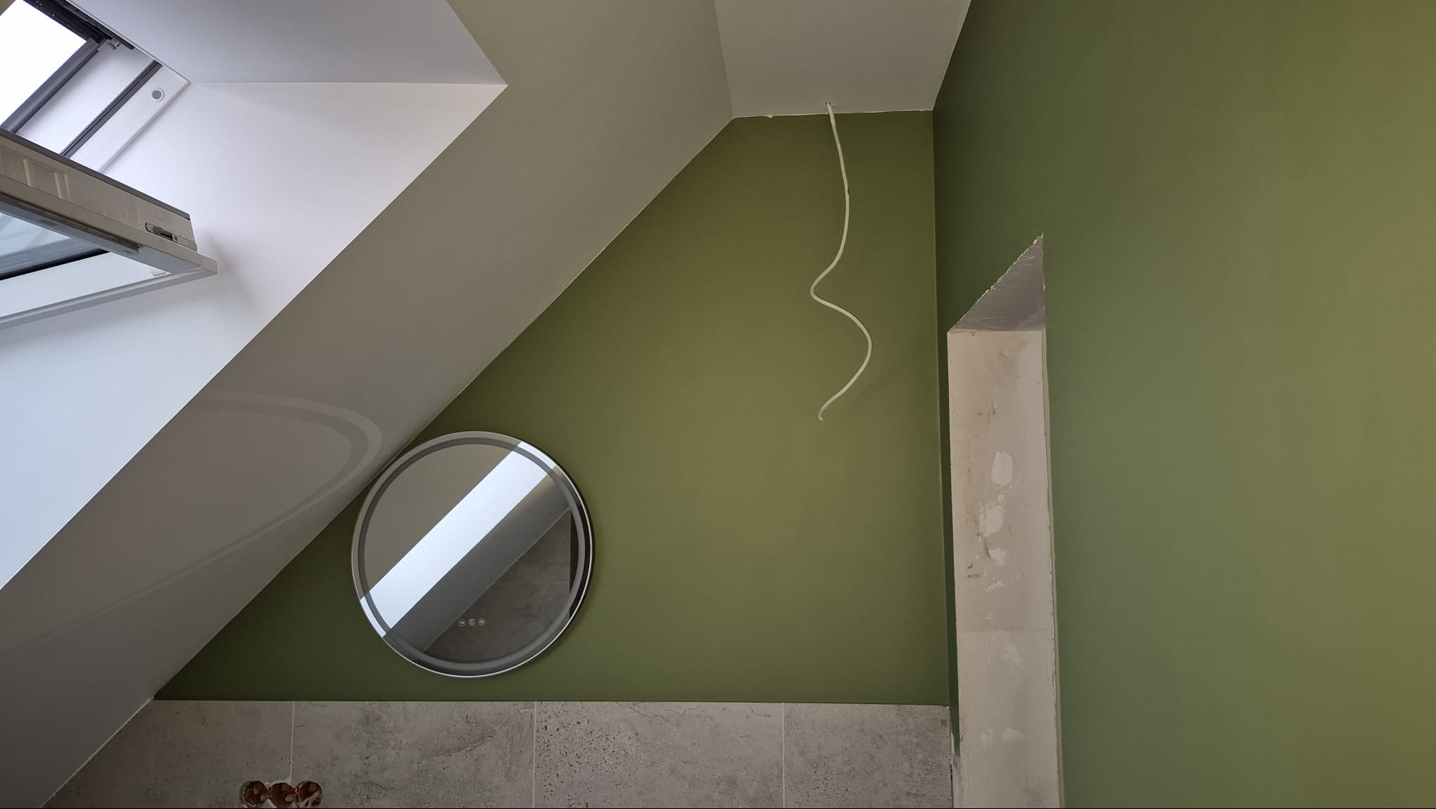 Ściana w odcieniach zieleni w łazience na poddaszu, okrągłe lustro z podświetleniem, widoczne przewody i fragment obudowy. Ukośne okno dachowe.