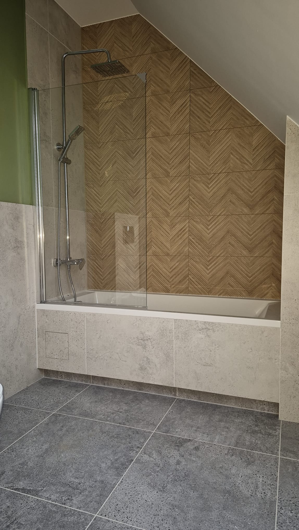 Wykończona łazienka na poddaszu: szare płytki podłogowe i ścienne, kabina prysznicowa z deszczownicą, ściana z płytek imitujących drewno ułożonych w jodełkę.