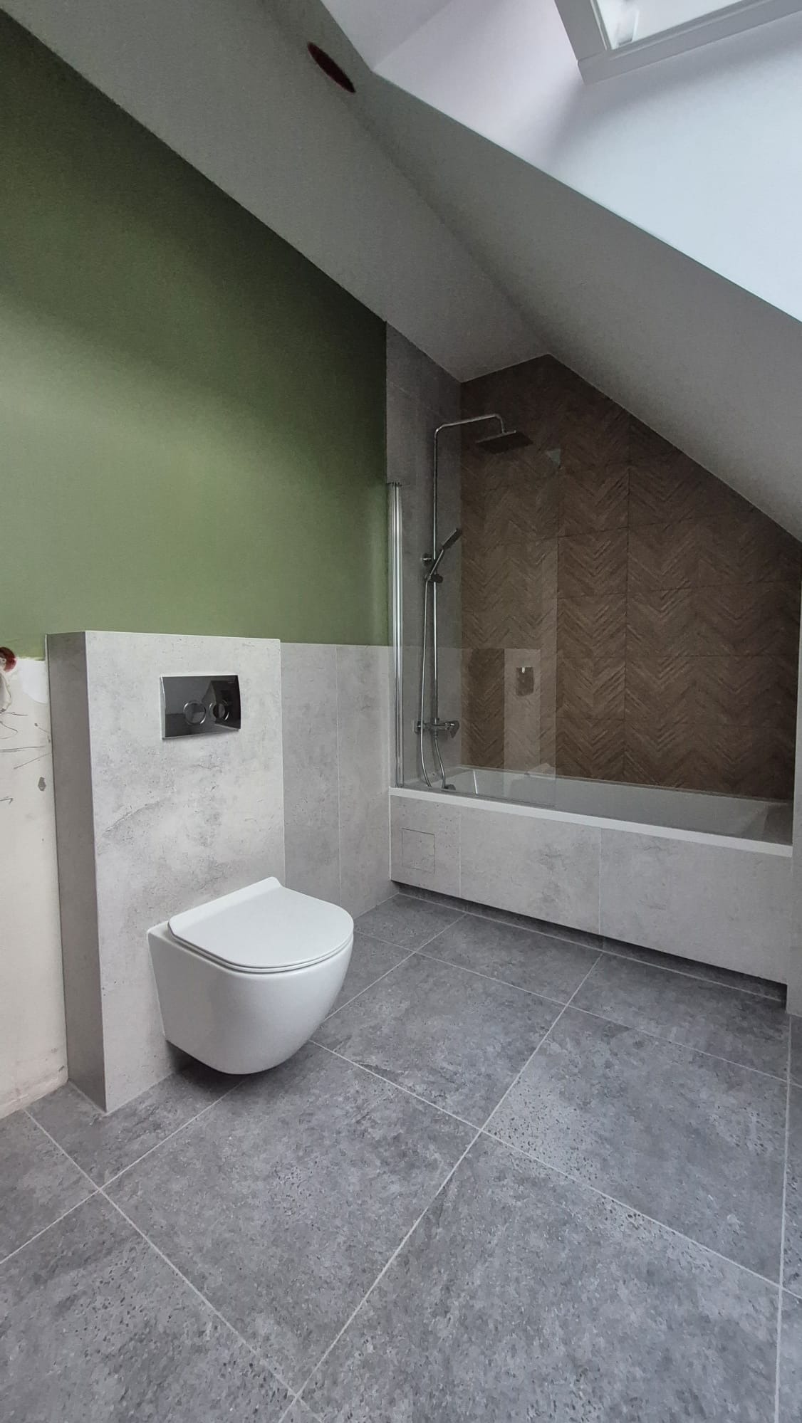 Wykończona łazienka na poddaszu z szarą podłogą i ścianami. Wisząca toaleta, kabina prysznicowa za szklaną ścianką. Jedna ściana pomalowana na zielono.