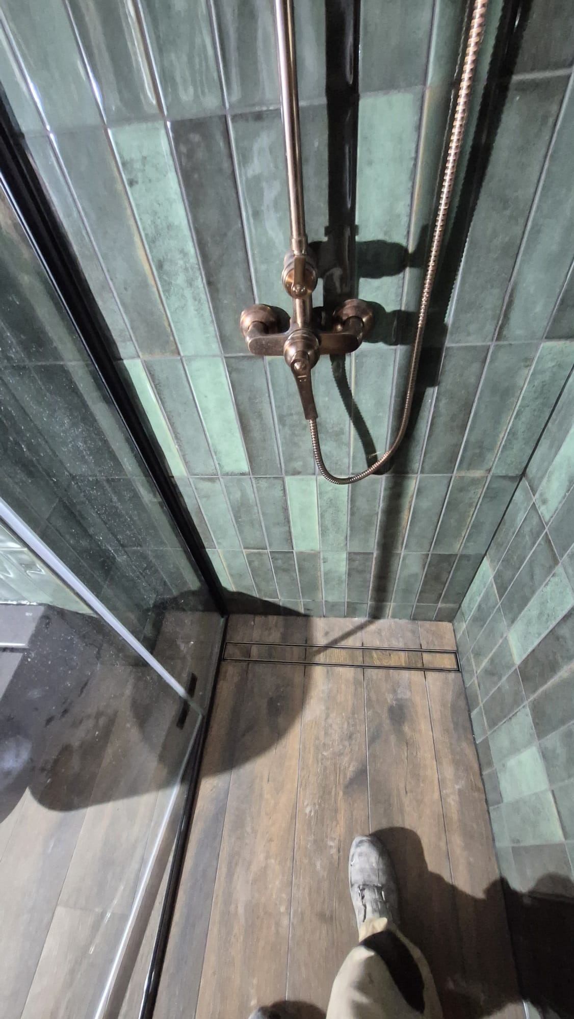 Kabina prysznicowa z zielonymi, podłużnymi płytkami i miedzianą armaturą. Widoczny odpływ liniowy i fragment buta. Ujęcie z góry.
