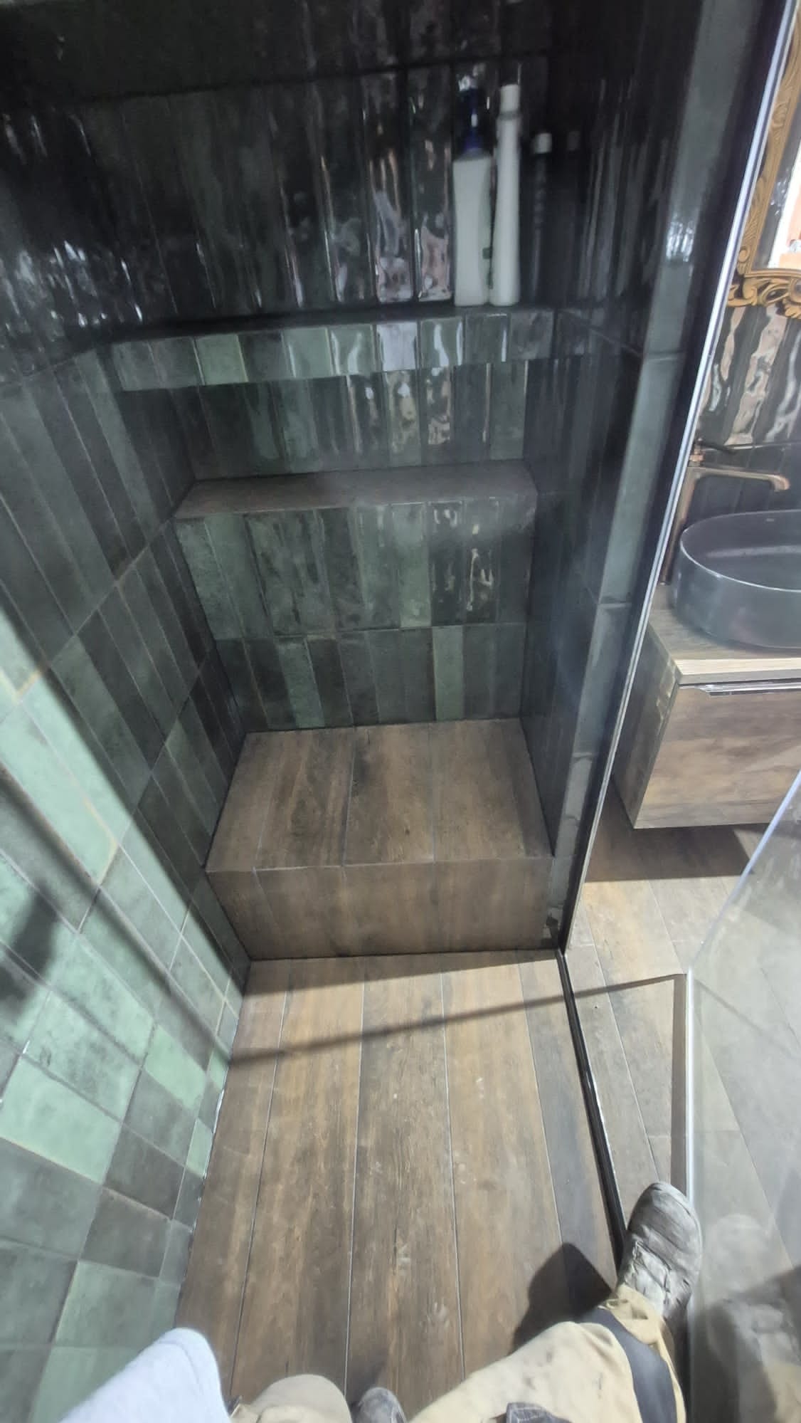 Wykończona kabina prysznicowa z ciemnozielonymi, błyszczącymi płytkami ułożonymi wertykalnie. Widoczna półka wewnątrz i fragment umywalki z szafką. Ujęcie z dołu.