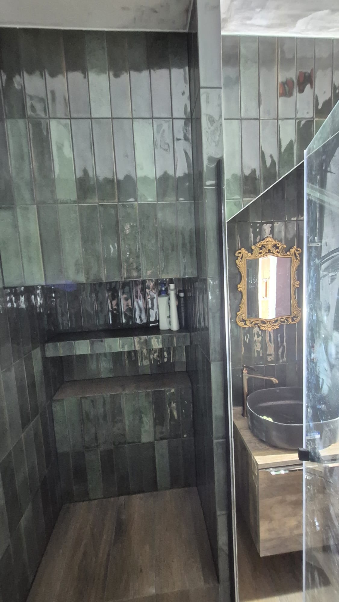 Nowoczesna kabina prysznicowa z ciemnozielonymi, błyszczącymi płytkami ułożonymi pionowo, drewnianymi półkami i umywalką z ozdobnym lustrem w złotej ramie.