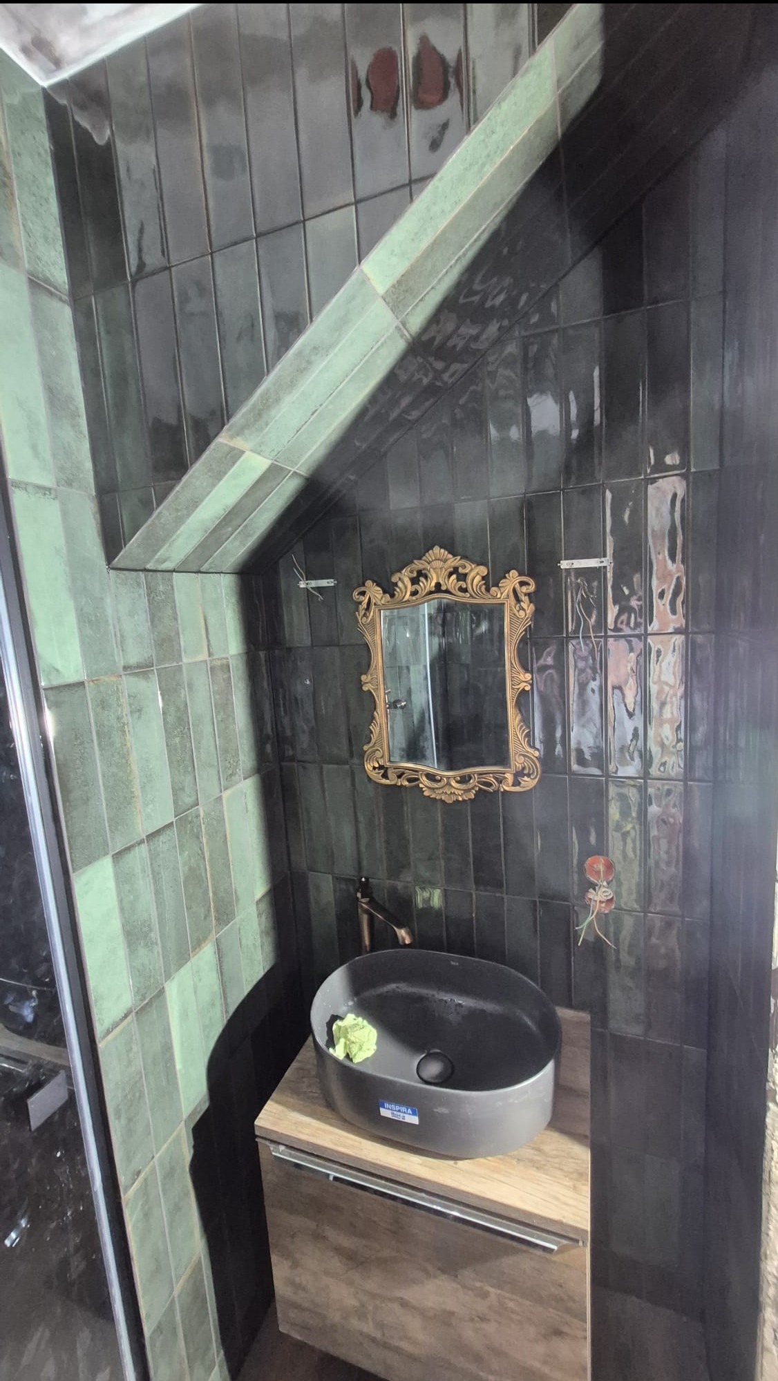 Wykończona łazienka z ciemnozielonymi, błyszczącymi płytkami ułożonymi pionowo, umywalką nablatową i lustrem w złotej, ozdobnej ramie. Nietypowe ułożenie płytek pod skosem.