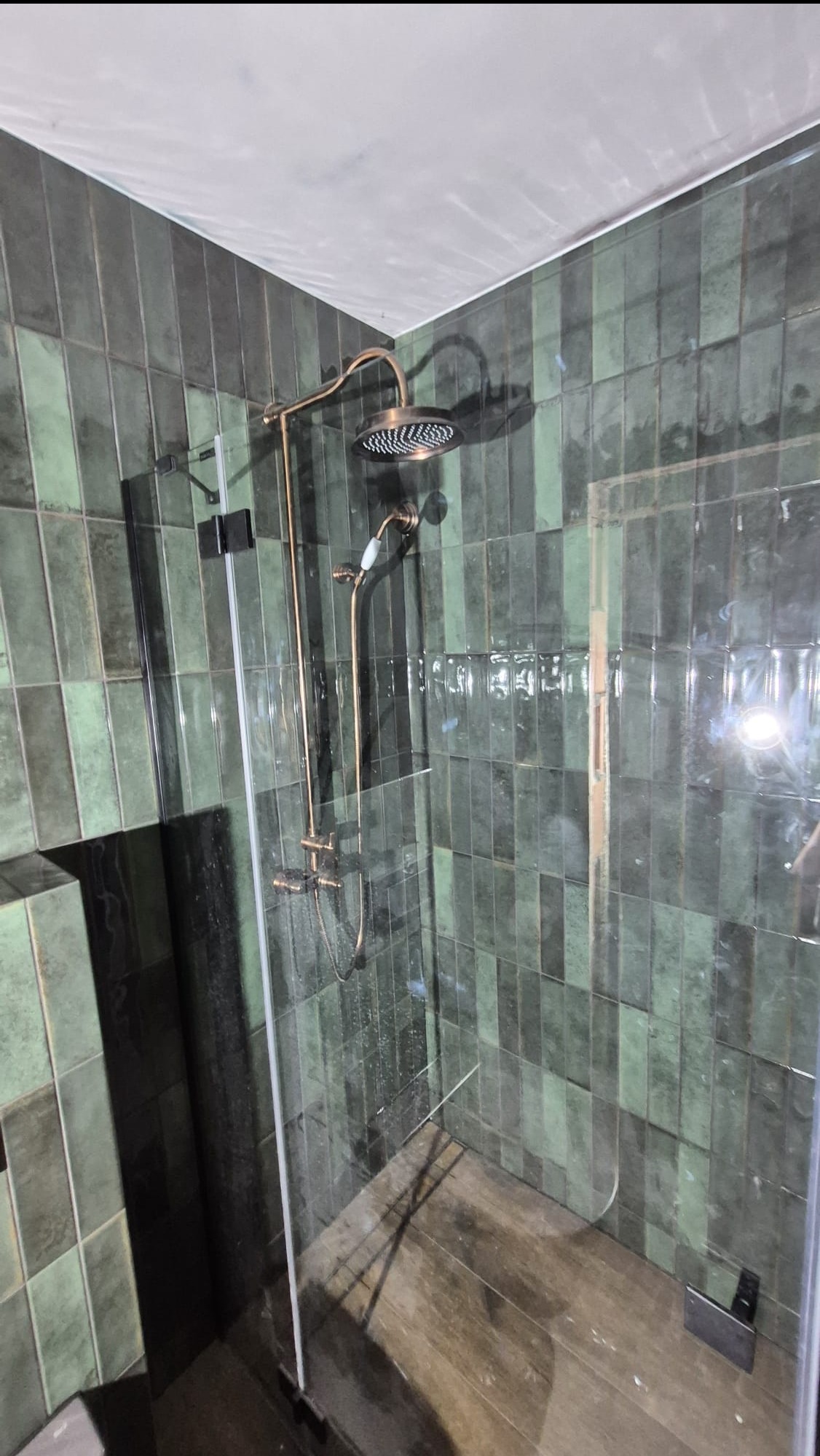 Kabina prysznicowa z zielonymi, pionowo ułożonymi płytkami i złotą armaturą. Widoczne szklane drzwi i drewnopodobna podłoga. Ujęcie z dołu.