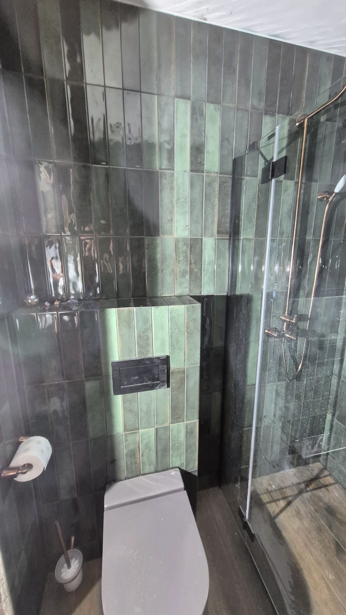 Nowoczesna łazienka z zielonymi, błyszczącymi płytkami ułożonymi pionowo, kabiną prysznicową ze złotymi elementami i podwieszaną toaletą.