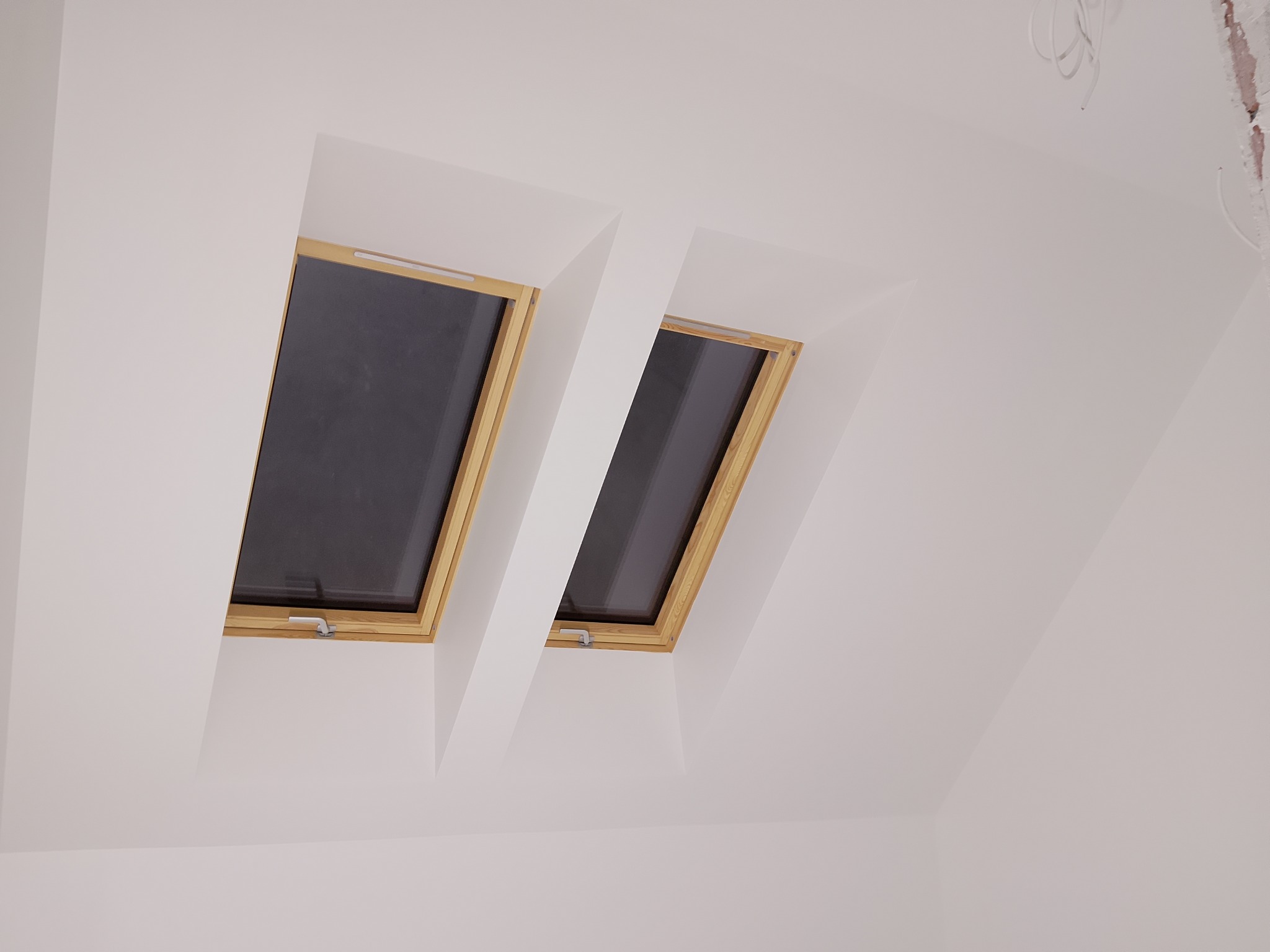 Dwa okna dachowe z drewnianymi ramami, osadzone w białej, skośnej ścianie poddasza. Widoczne przewody elektryczne. Minimalistyczny design, jasne wnętrze.