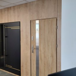 MB SALON OKIEN I DRZWI Monika Bronk - Korytarz biurowy z trzema parami drzwi: szklane, czarne z złotymi paskami i drewniane z numerem 12, zegar na ścianie. Nowoczesne wykończenie.