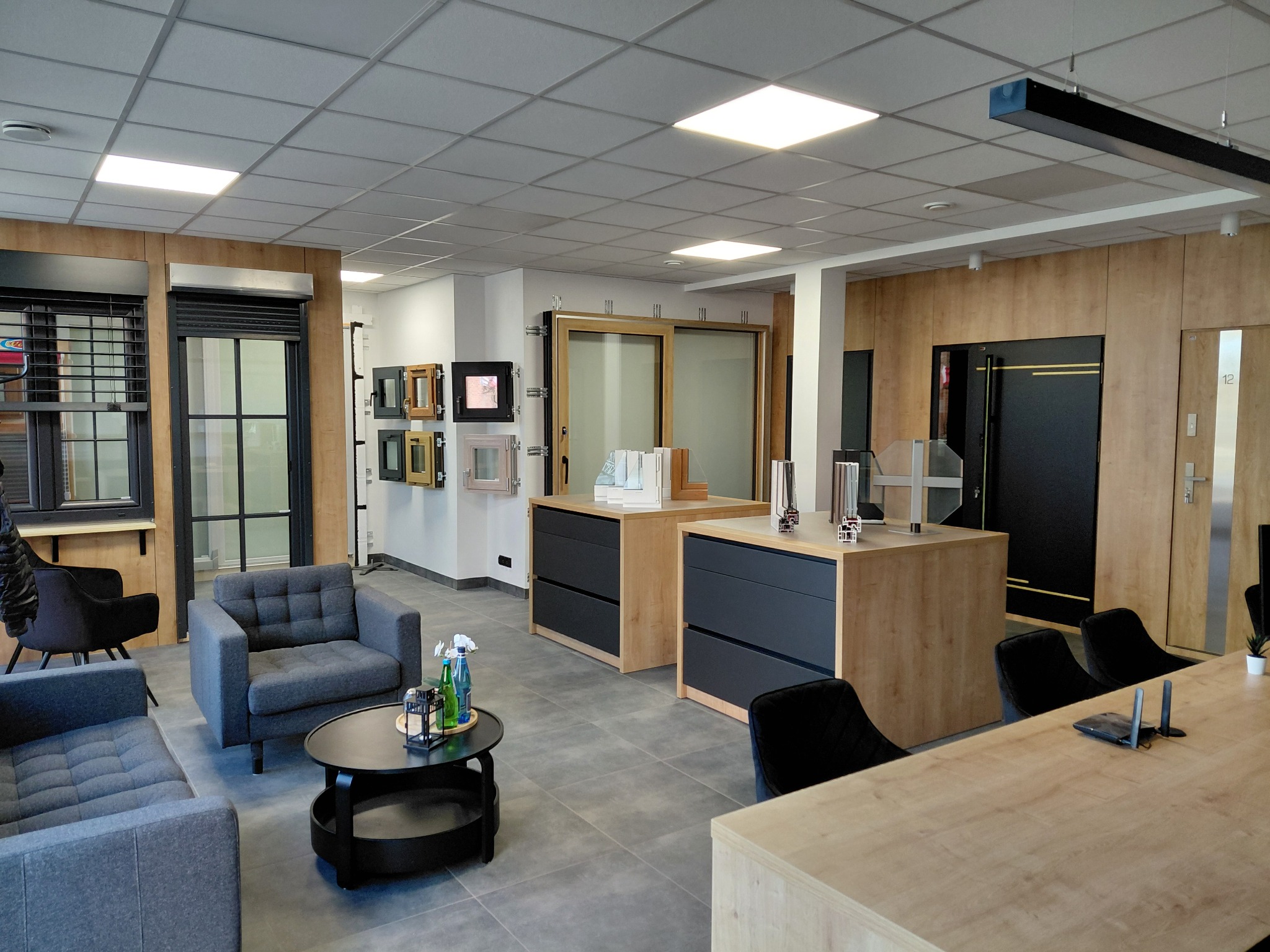 Ekspozycja w salonie: okna z roletami, drzwi przesuwne i antywłamaniowe. Aranżacja z sofami i stolikiem kawowym. Jasne oświetlenie, drewniane wykończenia ścian.