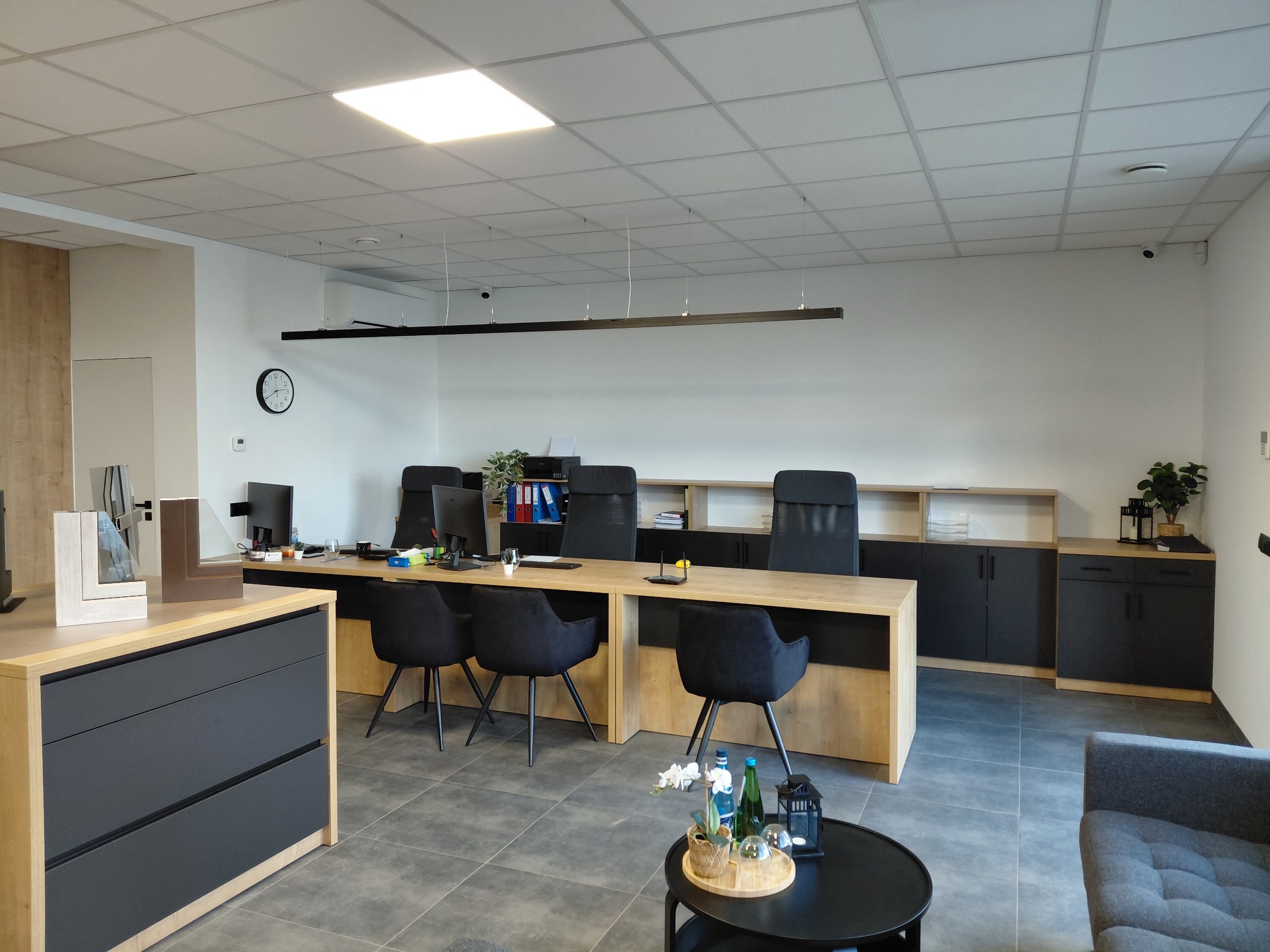Nowoczesne biuro z dużym stołem, czarnymi krzesłami i szafkami. Na stole komputery, dokumenty i dekoracje. Widoczne próbki okien. Stonowana kolorystyka, minimalistyczny design.