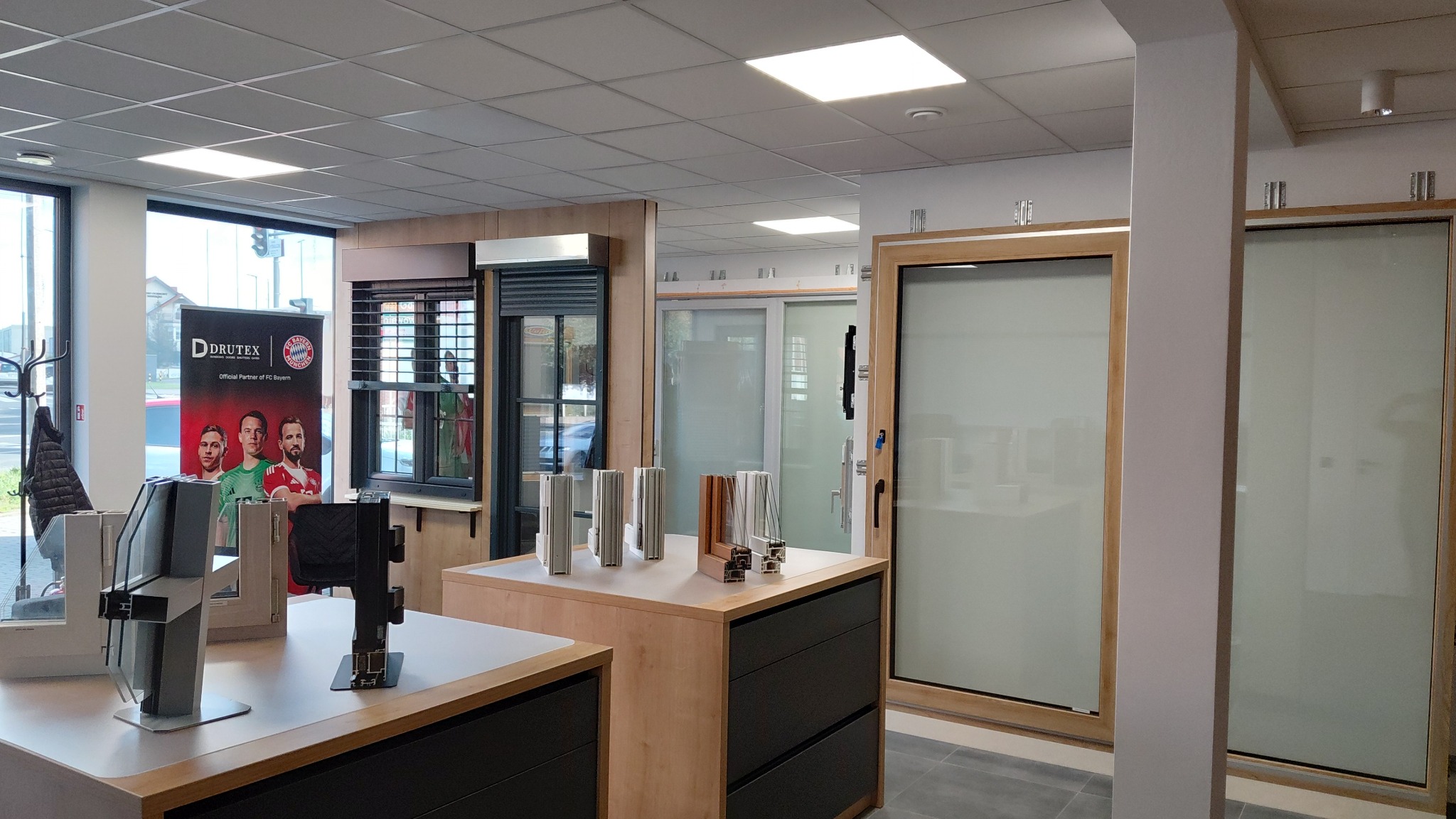 Salon z ekspozycją okien i drzwi: rolety zewnętrzne, okna PCV i aluminiowe, drzwi wewnętrzne przesuwne i szklane. W tle widoczny baner reklamowy.