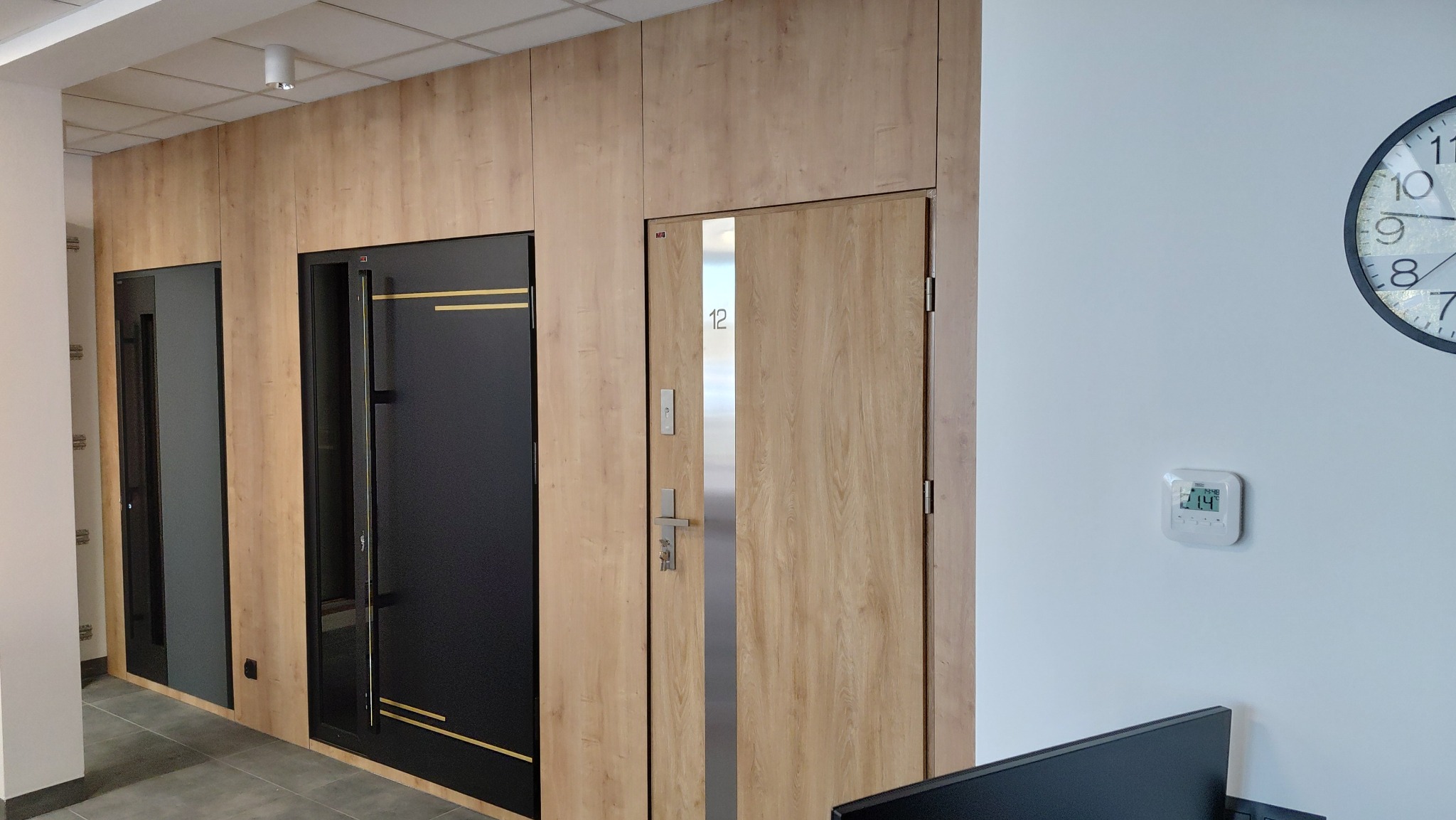 Korytarz biurowy z trzema parami drzwi: szklane, czarne z złotymi paskami i drewniane z numerem 12, zegar na ścianie. Nowoczesne wykończenie.