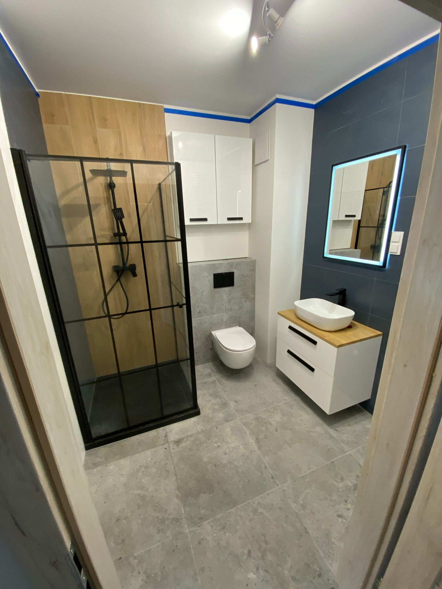 Nowoczesna łazienka po remoncie: kabina prysznicowa z czarną ramą, biała szafka z umywalką na drewnianym blacie i podświetlane lustro. Szare płytki na podłodze i ścianach.