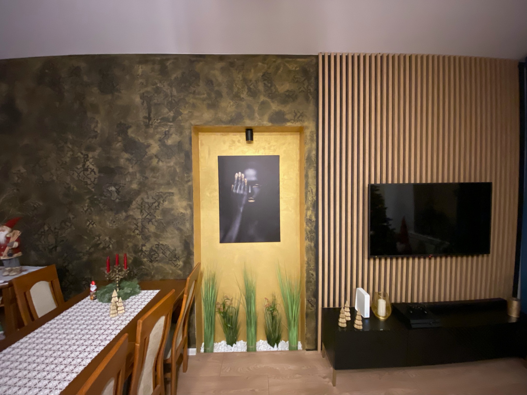 Stylowe wnętrze jadalni z dekoracyjną ścianą w odcieniach złota i brązu, drewnianymi lamelami, telewizorem i czarną komodą. Na stole świąteczne dekoracje.