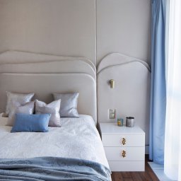 Joanna Lafuite-Tomaszewska we do design pl - Elegancka sypialnia z beżowym panelem tapicerowanym za łóżkiem, białą szafką nocną z ozdobnymi uchwytami i błękitnymi zasłonami. Parkiet z ciemnego drewna.