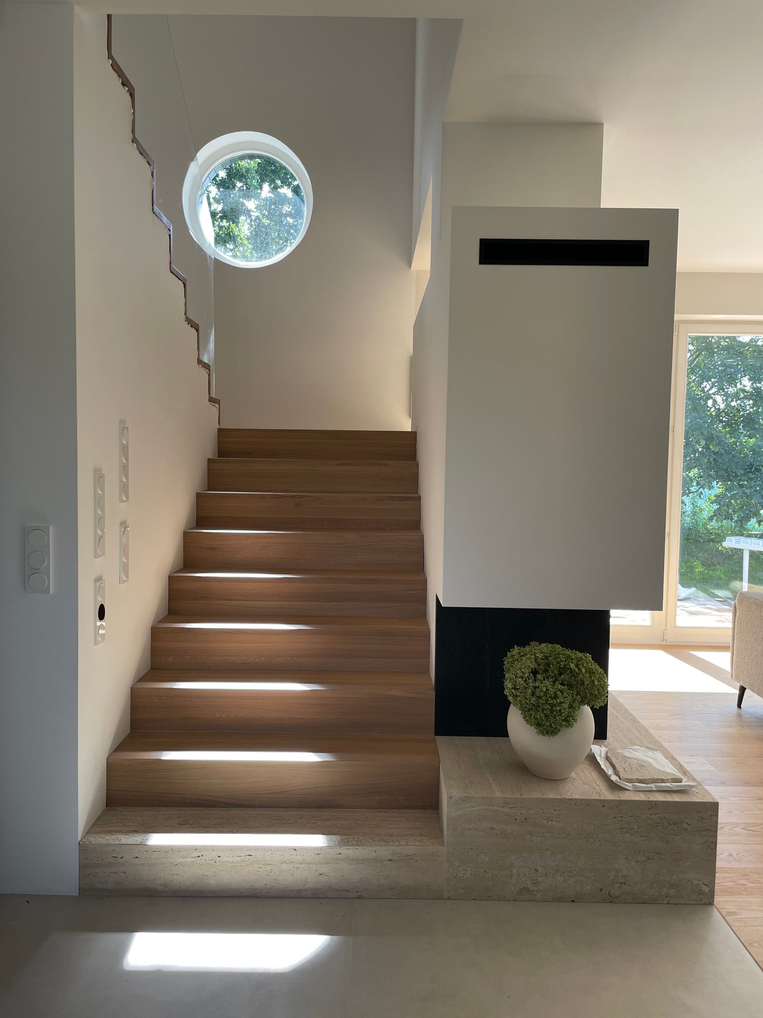 Nowoczesne wnętrze domu z drewnianymi schodami, okrągłym oknem i minimalistycznym kominkiem. Jasne, naturalne światło podkreśla detale aranżacji.