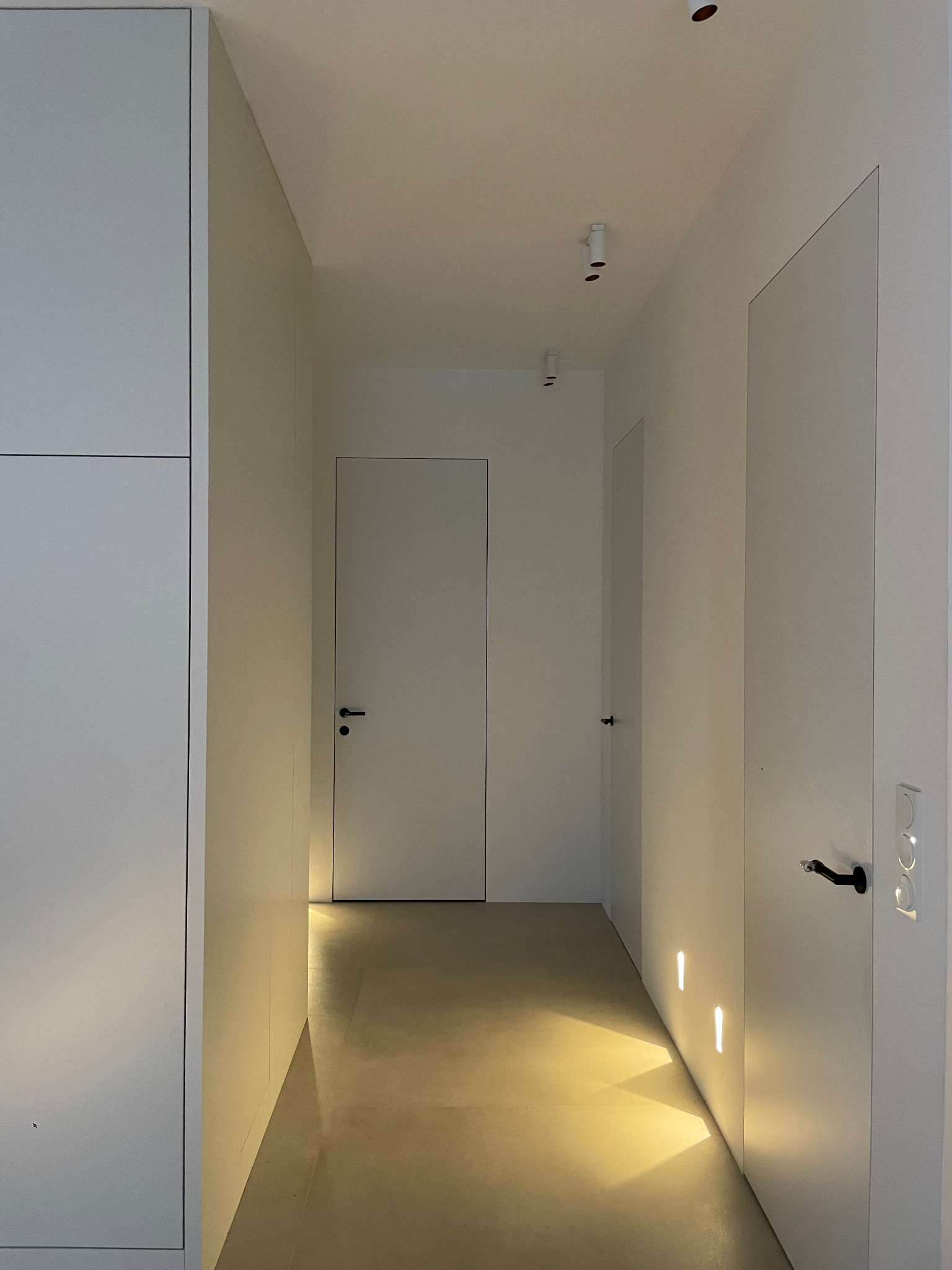 Minimalistyczny korytarz z białymi ścianami, drzwiami z czarnymi klamkami i subtelnym oświetleniem podłogowym. Widoczne szafy w zabudowie i punktowe lampy sufitowe.