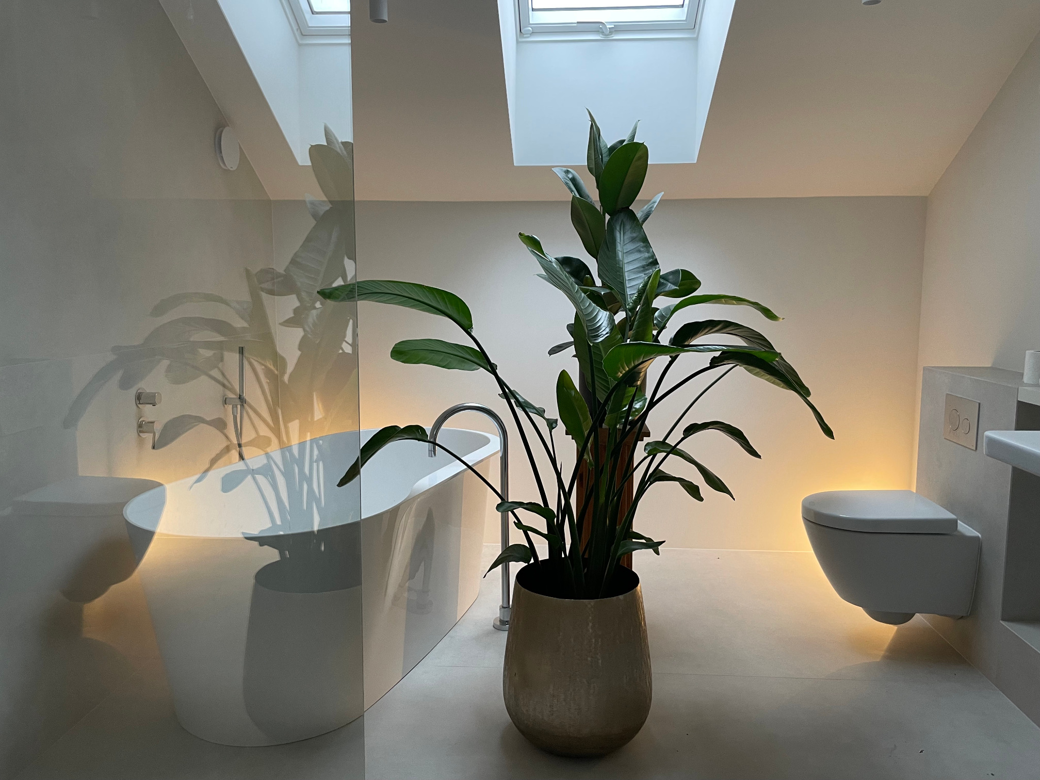 Jasna łazienka z wolnostojącą wanną za szklaną ścianką, toaletą podwieszaną z podświetleniem i dużą rośliną w donicy. Okno dachowe w suficie.