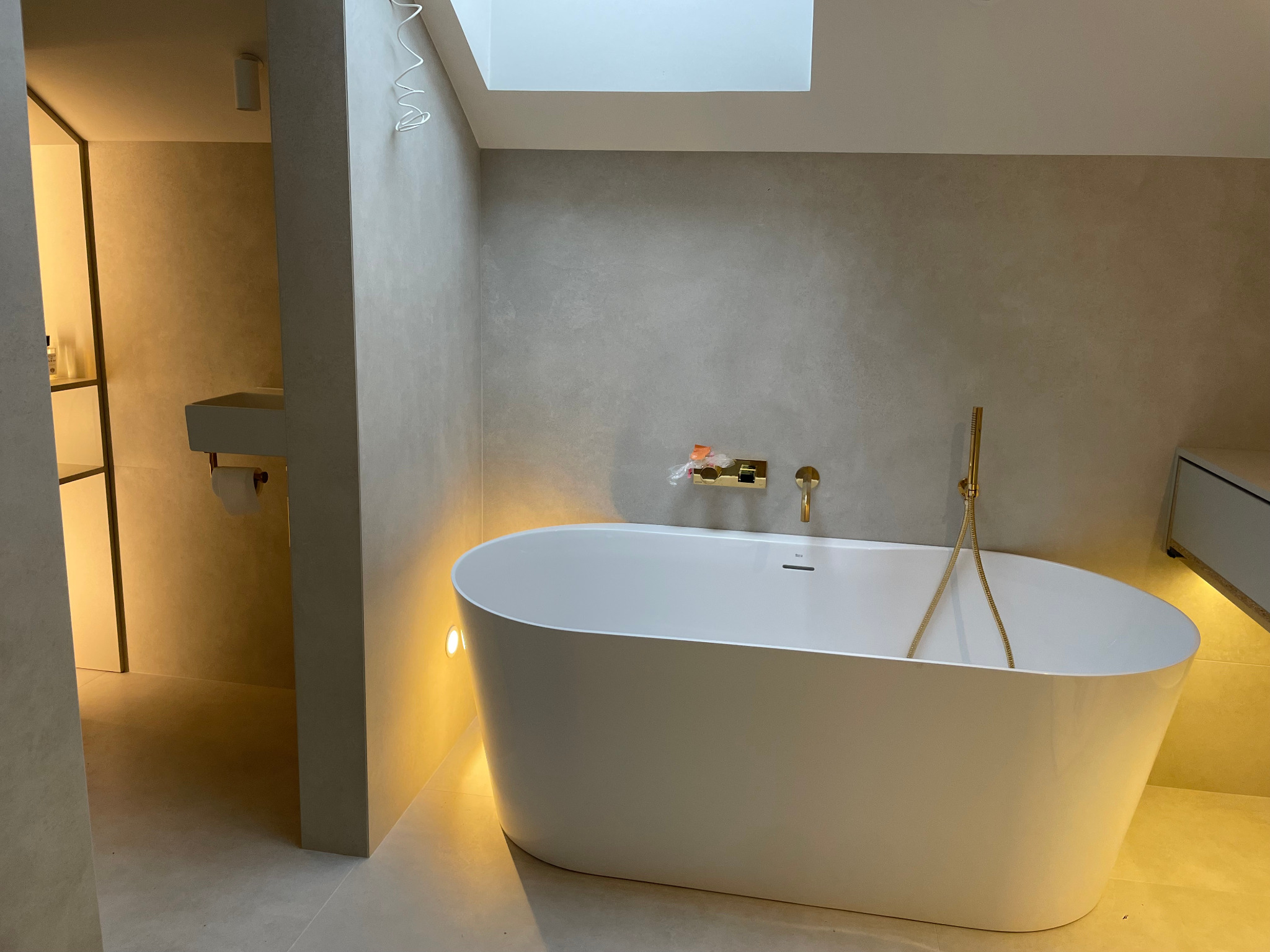 Nowoczesna łazienka z wolnostojącą białą wanną, złotą armaturą i subtelnym oświetleniem punktowym. Minimalistyczny design, jasne kolory i okno dachowe.