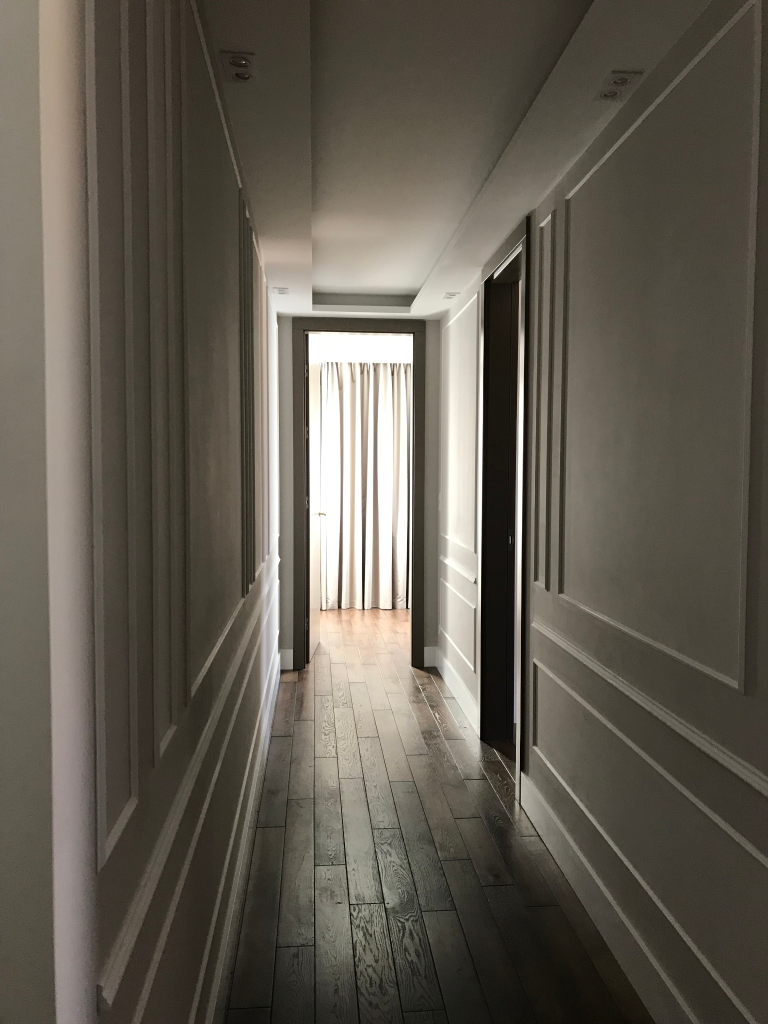 Elegancki korytarz z panelami ściennymi w odcieniach szarości i drewnianą podłogą. Widok w głąb, na końcu zasłonięte okno. Minimalistyczne oświetlenie sufitowe.