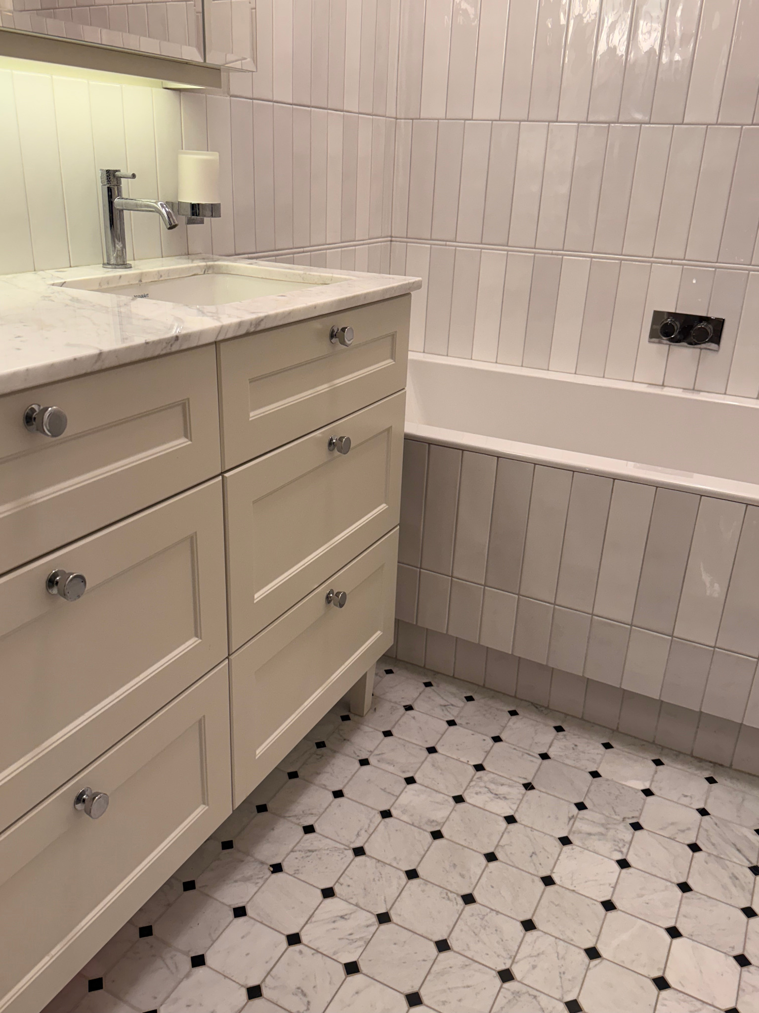 Jasna łazienka z marmurową umywalką, białymi płytkami ułożonymi pionowo i podłogą w biało-czarną szachownicę. Bateria umywalkowa chromowana, minimalistyczna.