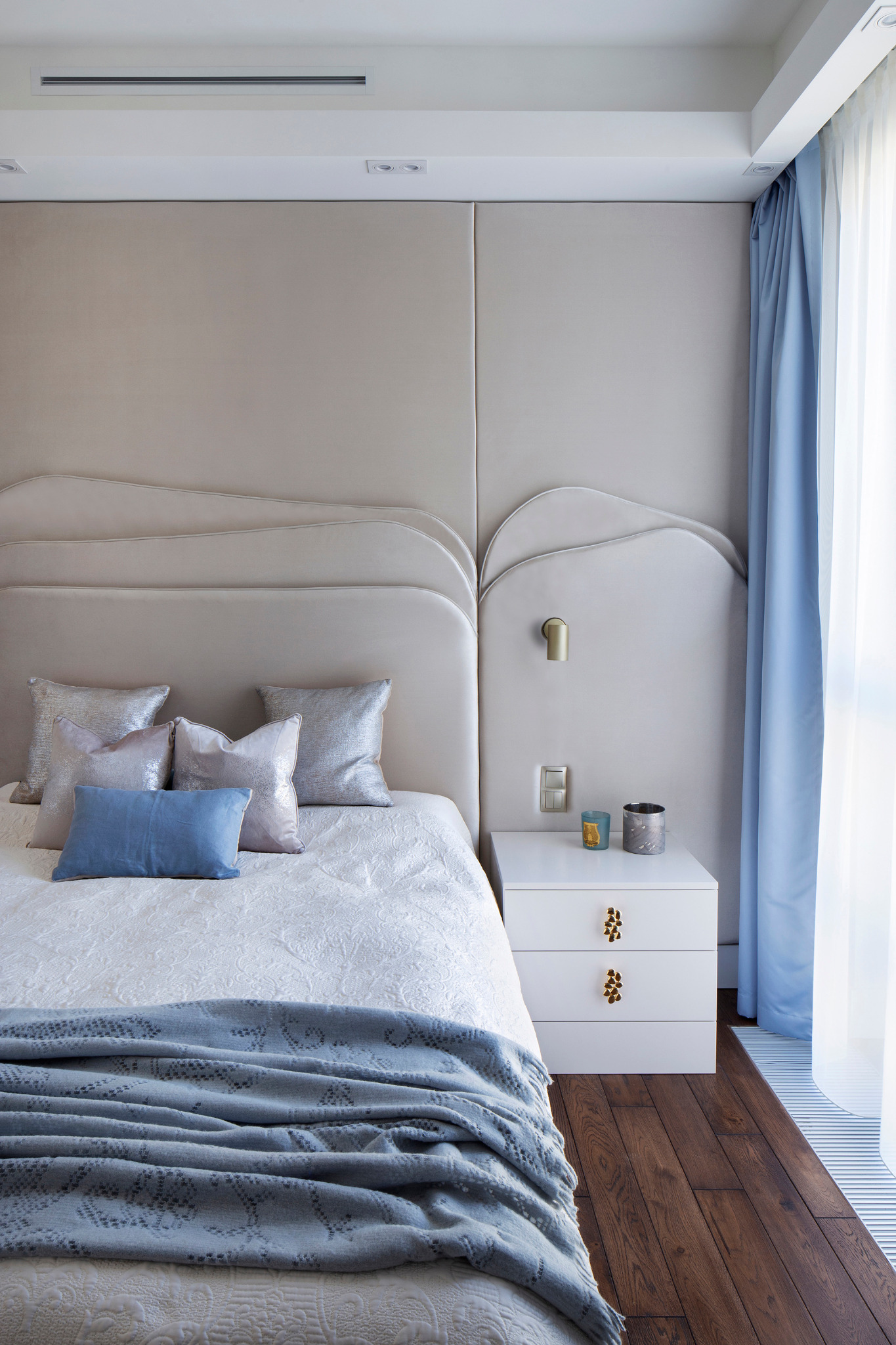 Elegancka sypialnia z beżowym panelem tapicerowanym za łóżkiem, białą szafką nocną z ozdobnymi uchwytami i błękitnymi zasłonami. Parkiet z ciemnego drewna.