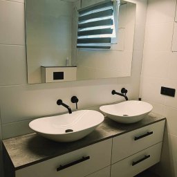 Daniel Kułakowski -Usługi i Projekty Remontowo-Techniczne - Nowoczesna łazienka z dwiema owalnymi umywalkami nablatowymi i czarną armaturą. Białe płytki na ścianach, lustro z podświetleniem LED i szafka z szufladami.