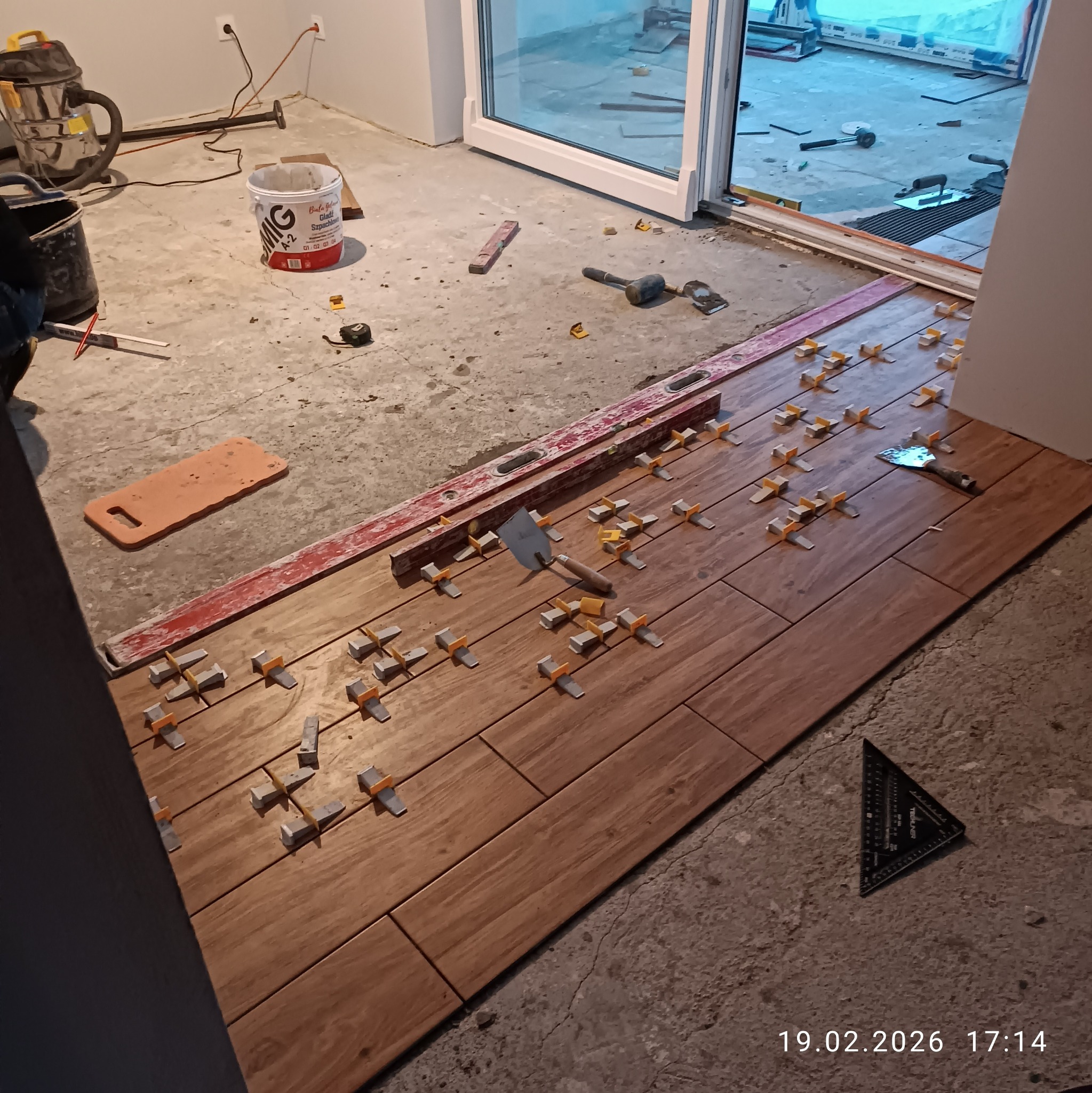 Układanie brązowych płytek imitujących drewno na podłodze, z użyciem poziomicy, kielni i krzyżyków dystansowych. Widoczne narzędzia i materiały budowlane.