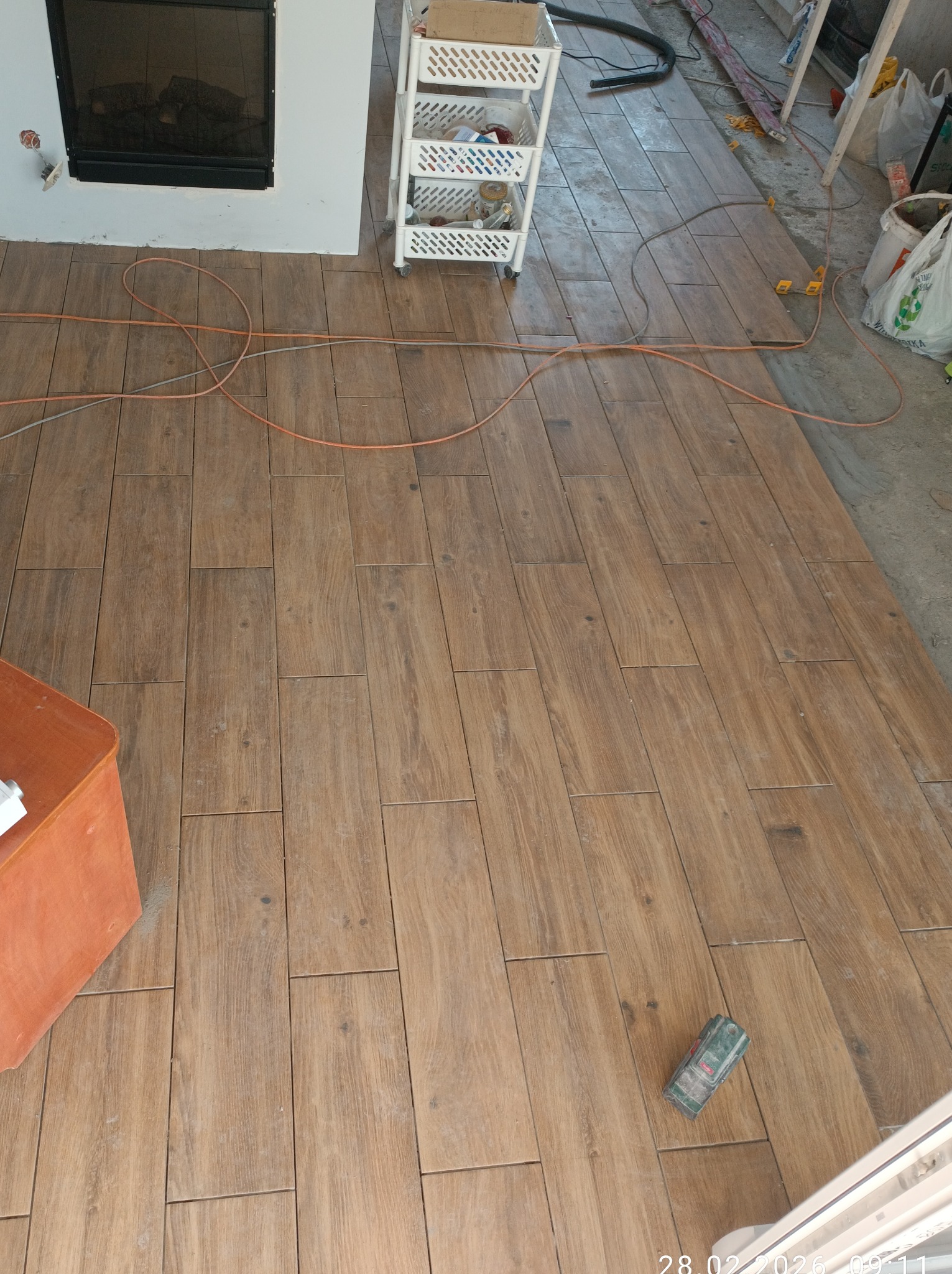 Ułożona podłoga z płytek imitujących drewno, widoczne kable, poziomica i akcesoria. Fragment kominka i regału na kółkach w tle. Układanie płytek podłogowych.