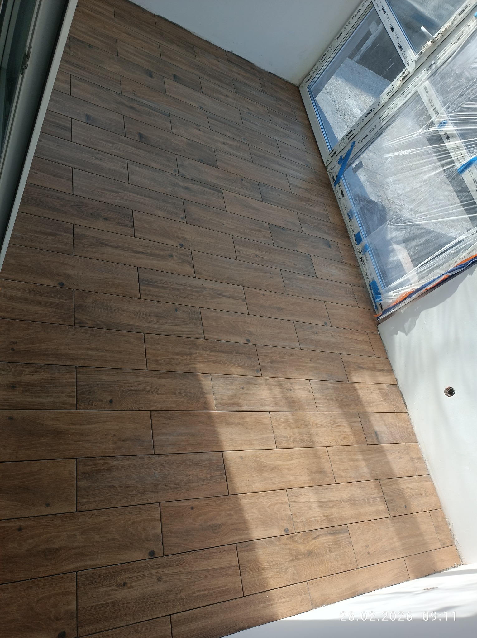 Ukończona podłoga z płytek imitujących drewno na balkonie, widok z góry, okno zabezpieczone folią. Ciemne płytki ułożone w regularny wzór, jasne ściany.