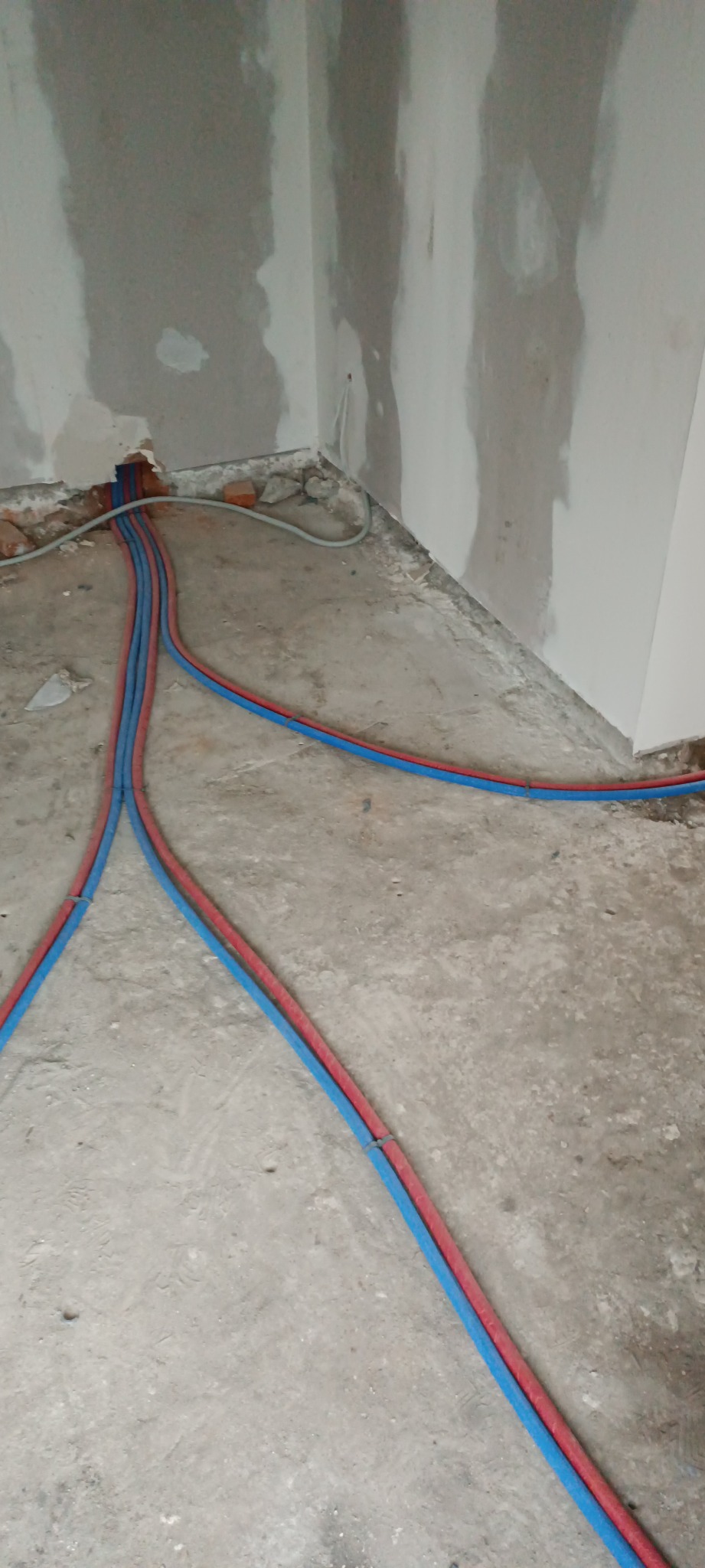 Fragment wnętrza w trakcie prac wykończeniowych: ściany z płyt gipsowych, na podłodze rury instalacji wodnej w kolorach czerwonym i niebieskim, wychodzące ze ściany.