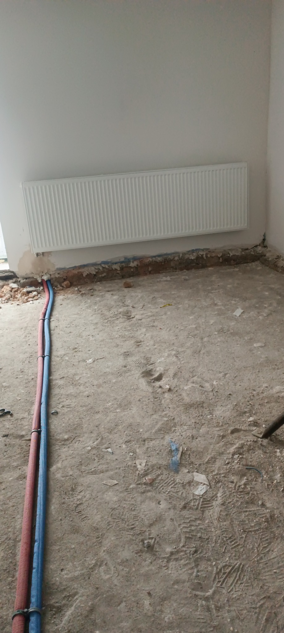 Biały grzejnik na ścianie, z widocznymi rurami instalacji grzewczej w podłodze w trakcie prac wykończeniowych. Surowy beton na podłodze.