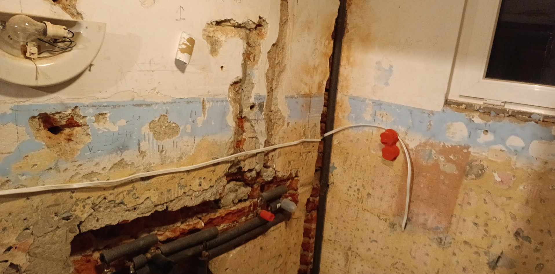 Zniszczona ściana w trakcie remontu z odsłoniętą instalacją elektryczną i rurami. Widoczne fragmenty starej farby i tynku, okno w prawym górnym rogu.