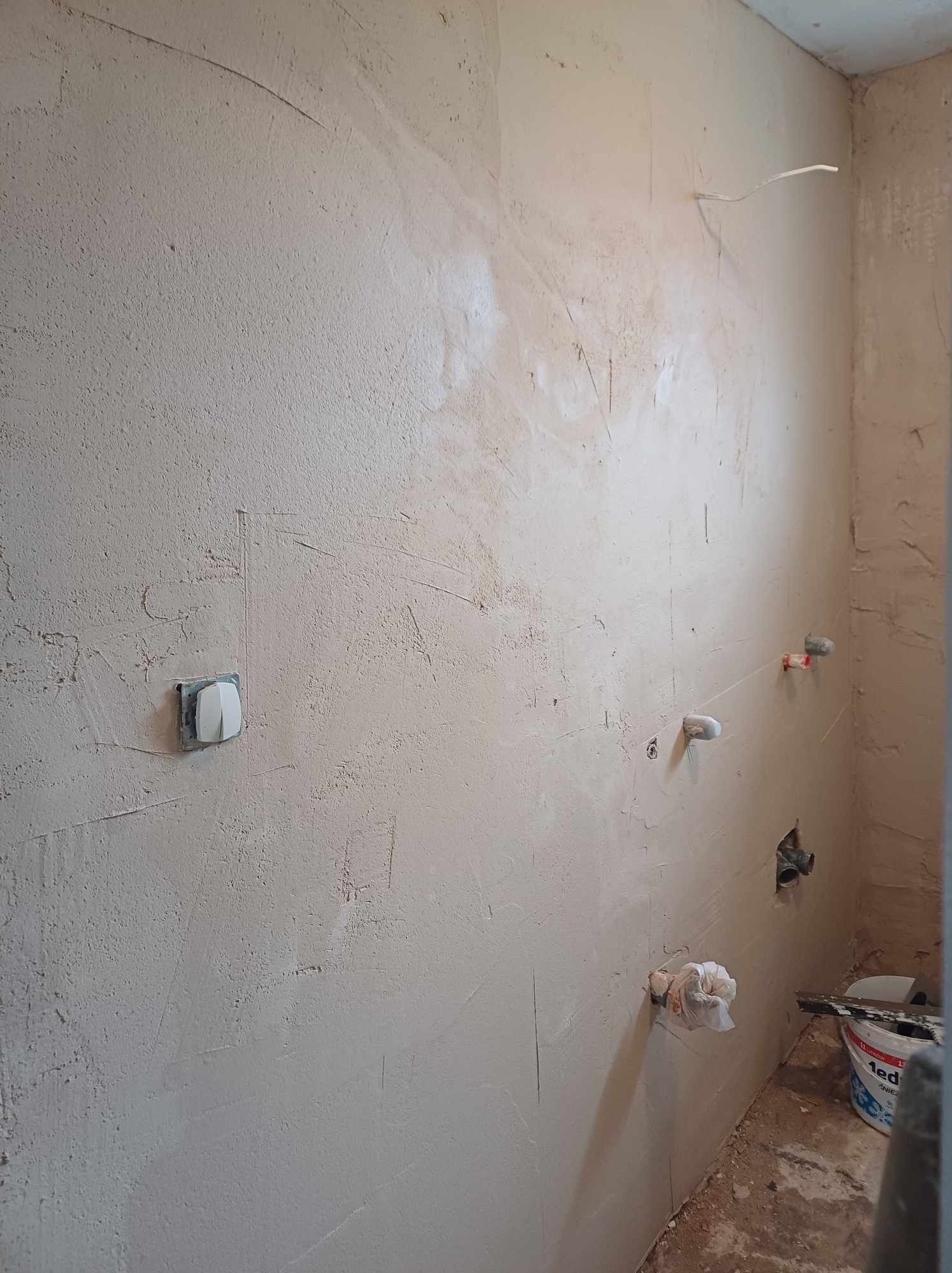 Ściana w trakcie gładzi w łazience z wystającymi rurami i puszką elektryczną. Widoczne narzędzia i wiadro z gładzią. Etap prac wykończeniowych.